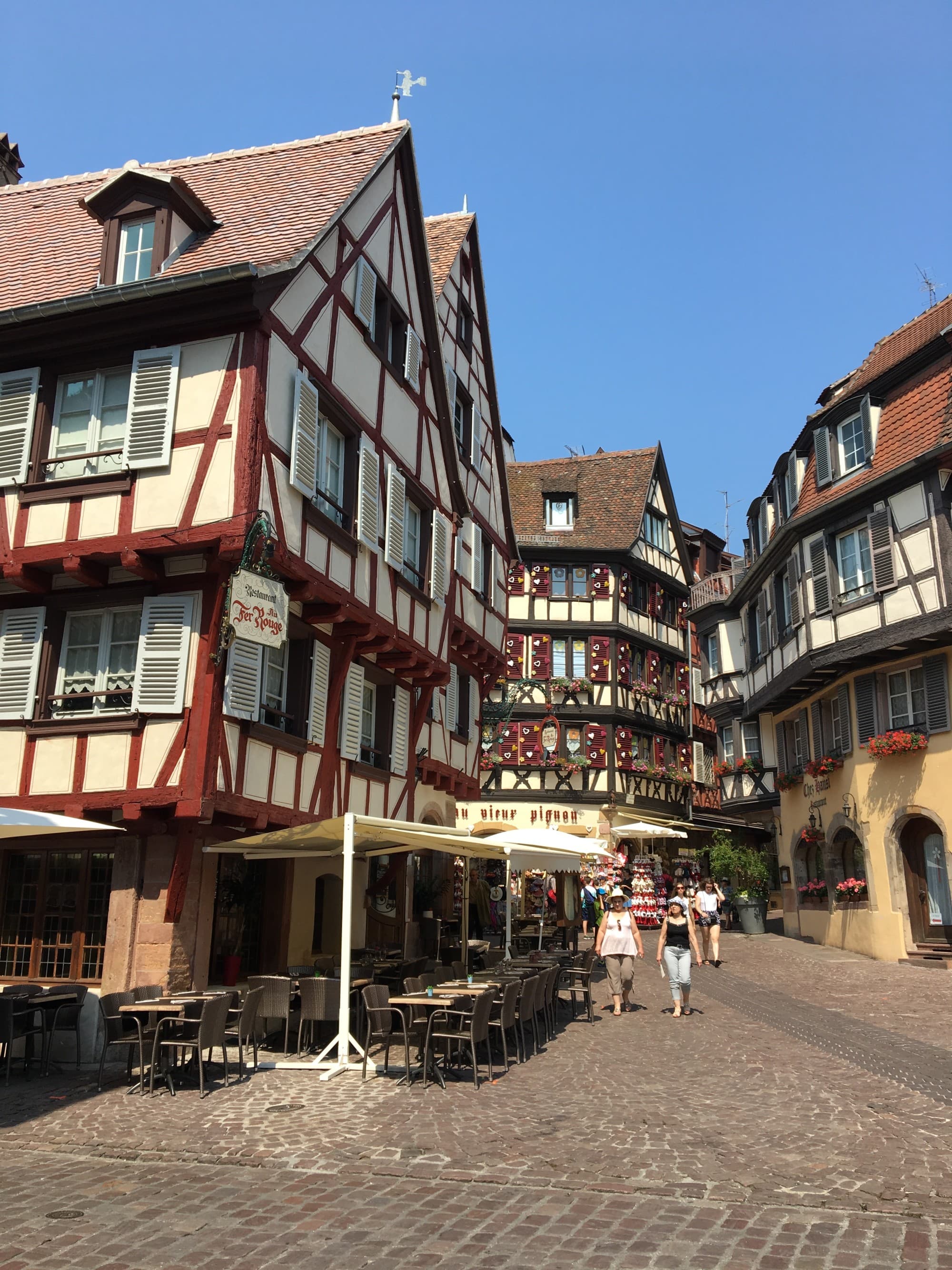 The image shows a quaint European street with half-timbered buildings and a clear blue sky.