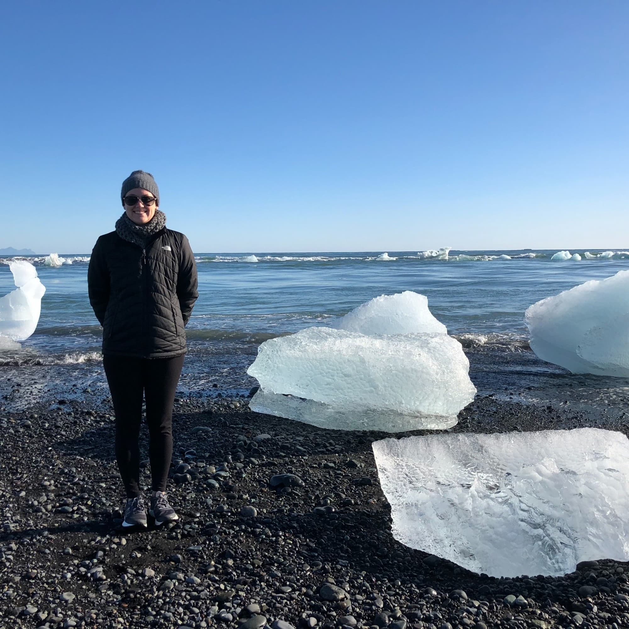 The image shows an individual standing on a pebble beach with large chunks of ice scattered around, against a backdrop of the ocean and a clear blue sky.