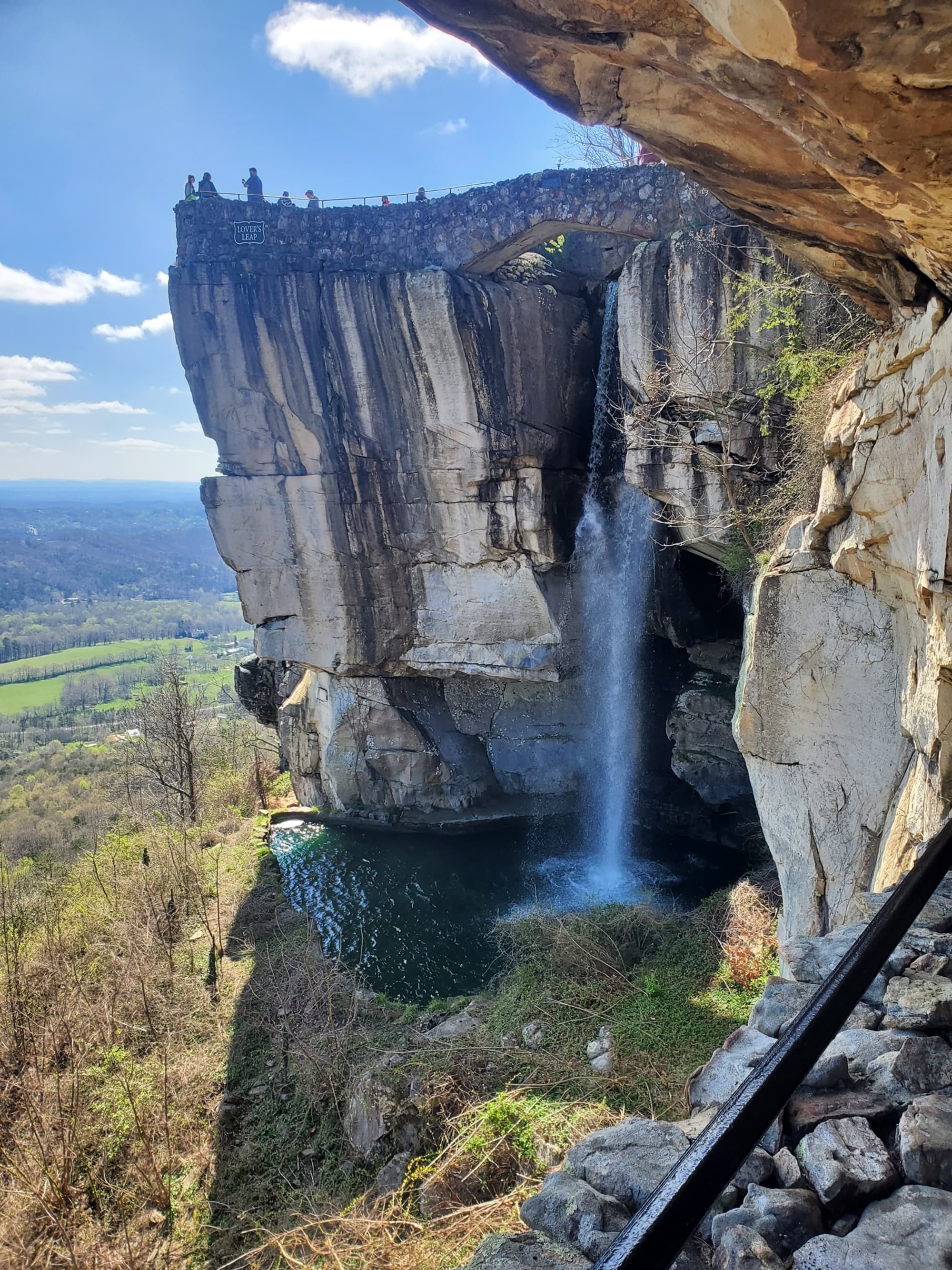 The image shows a waterfall cascading down from a rocky cliff into a pool below, surrounded by greenery and rock formations under a clear sky.