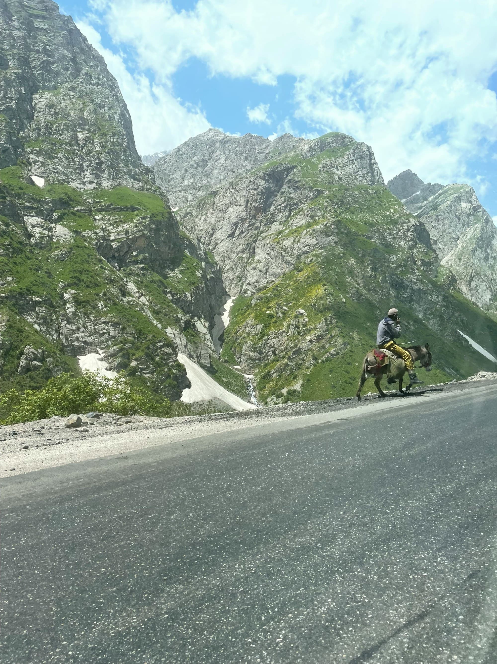 A person rides a horse on a road with towering, rugged mountains in the background.