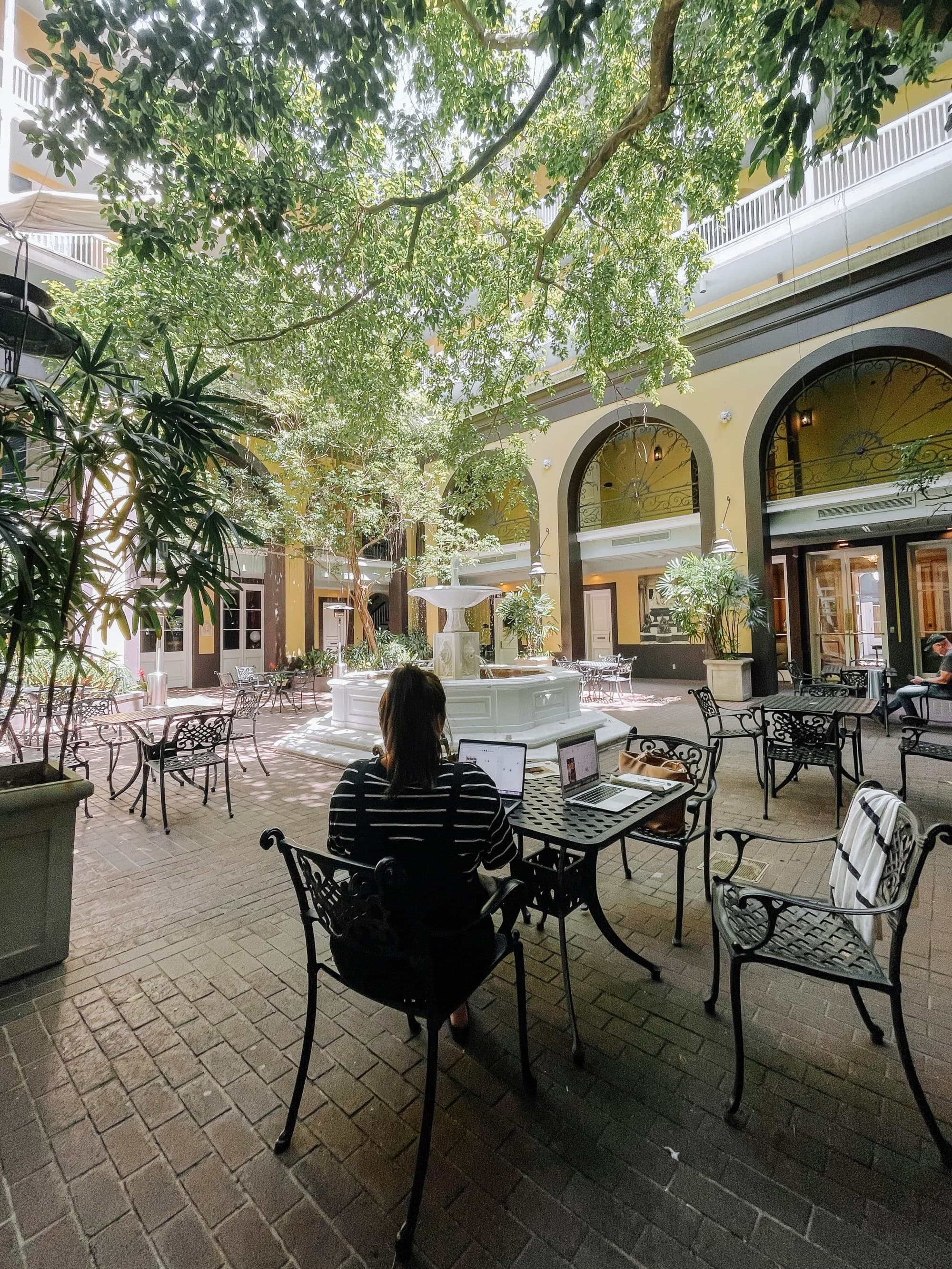 The image shows an open-air courtyard with greenery, seating areas, and a person working on a laptop.