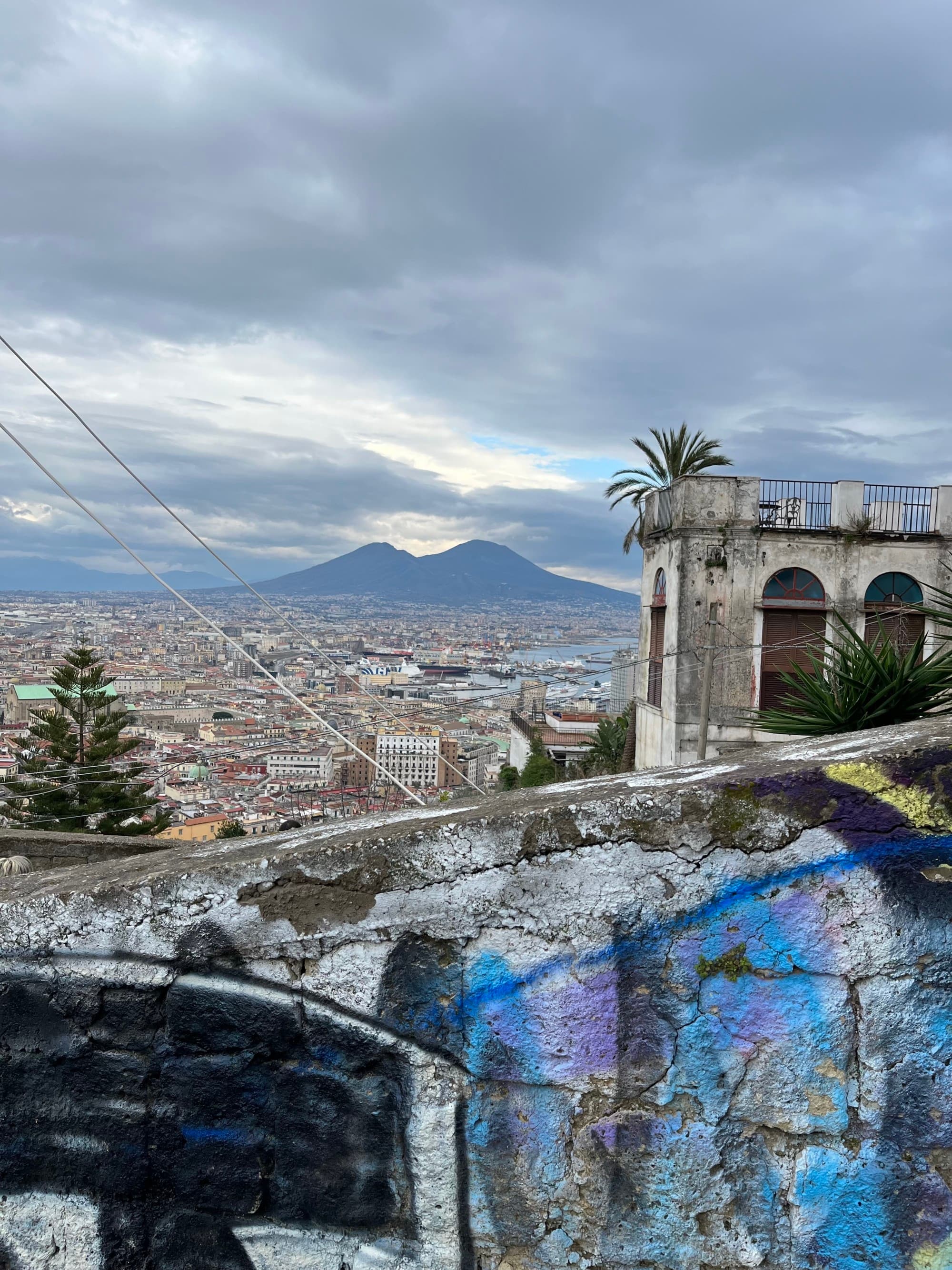 The image shows a panoramic view of a city with Mount Vesuvius in the background, taken from an elevated position with graffiti in the foreground.