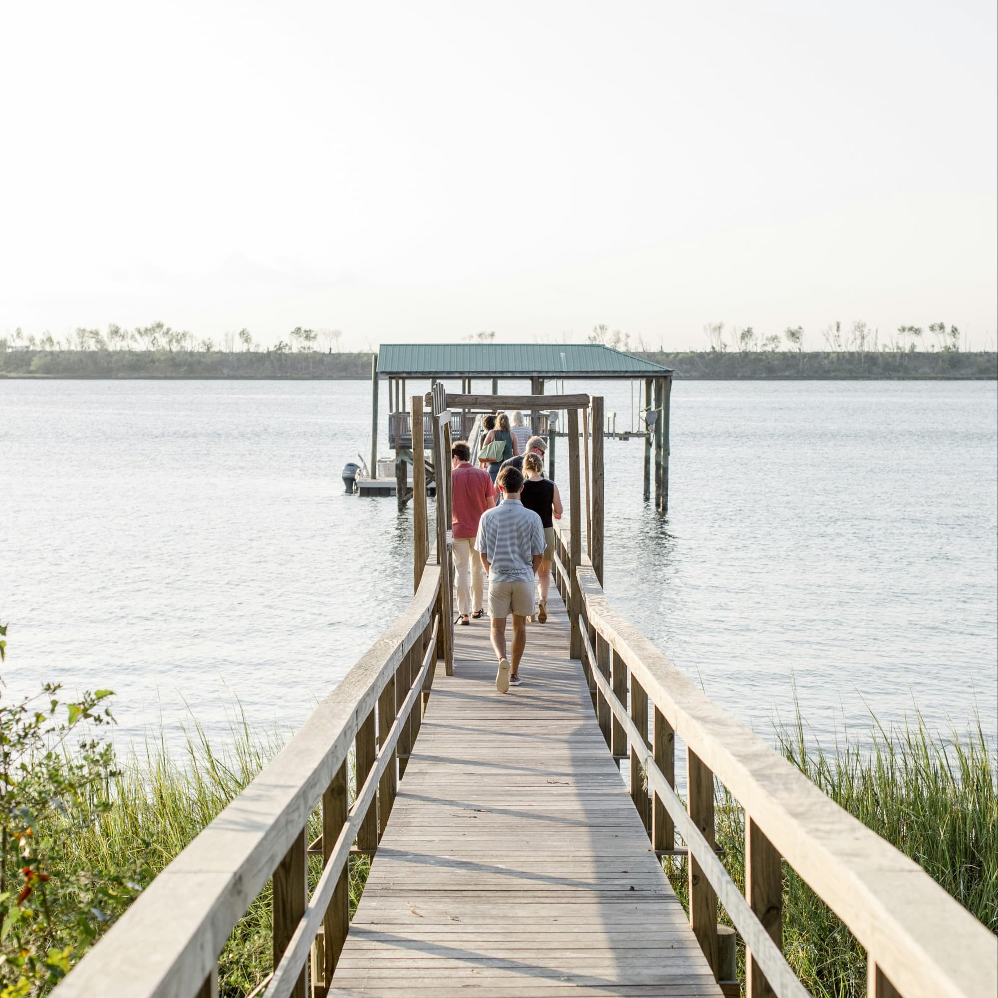 Three people are walking on a wooden pier towards a covered dock over a calm body of water, under a clear sky.