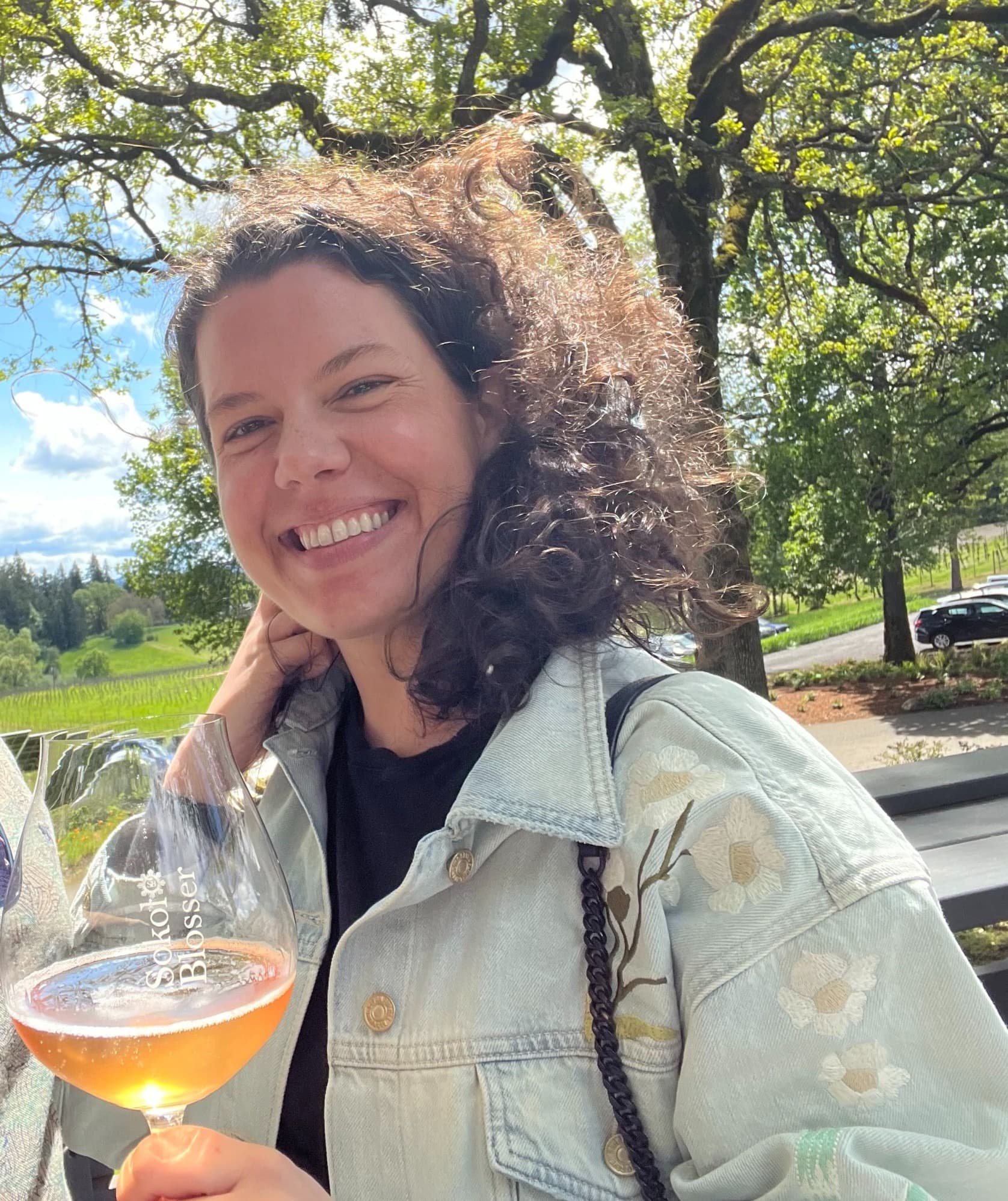 An individual holding a wine glass, with trees and a clear sky in the background.