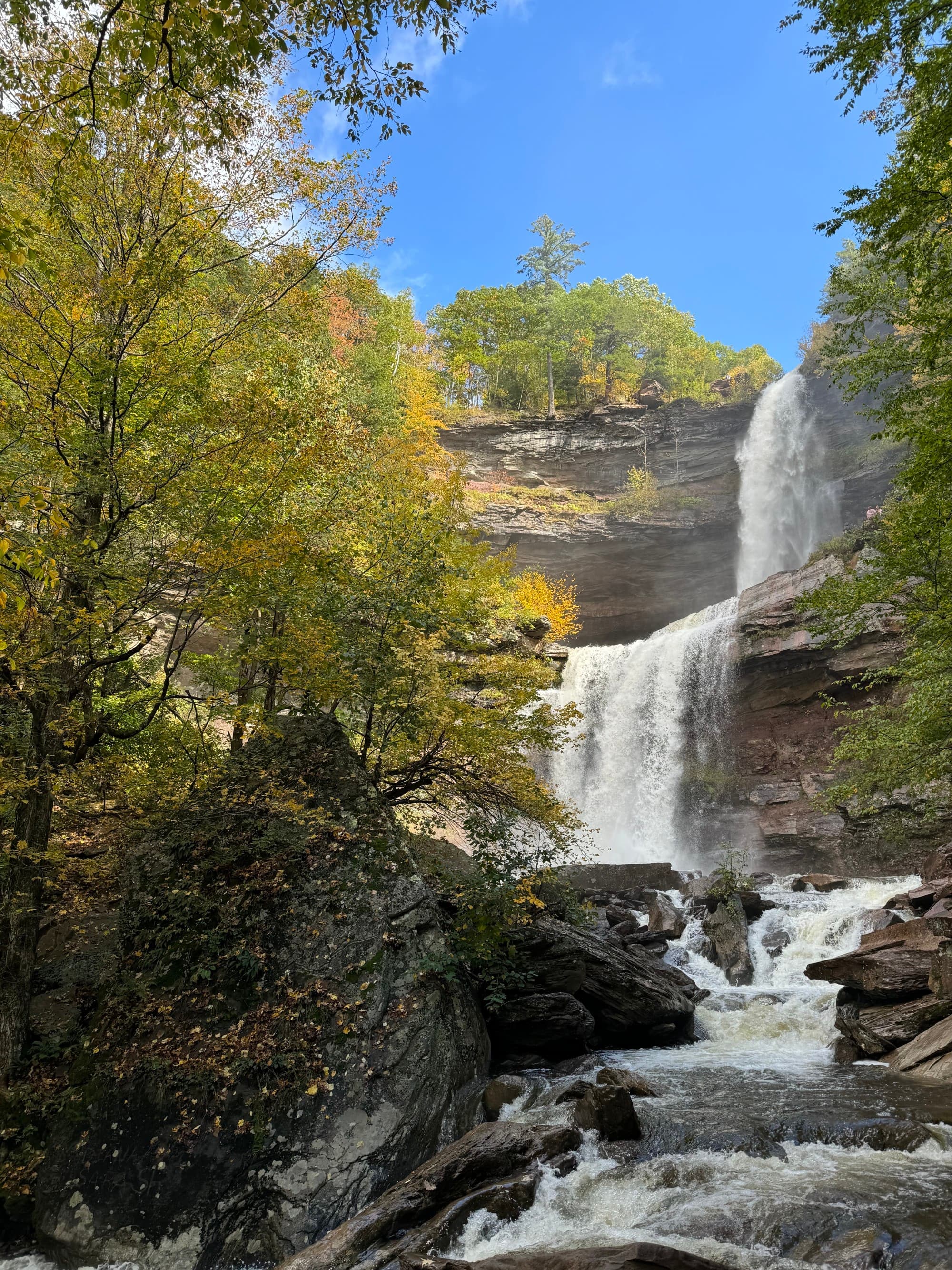 Kaaterskill Falls cascades down a rock face surrounded by lush, fall foliage on a sunny day.