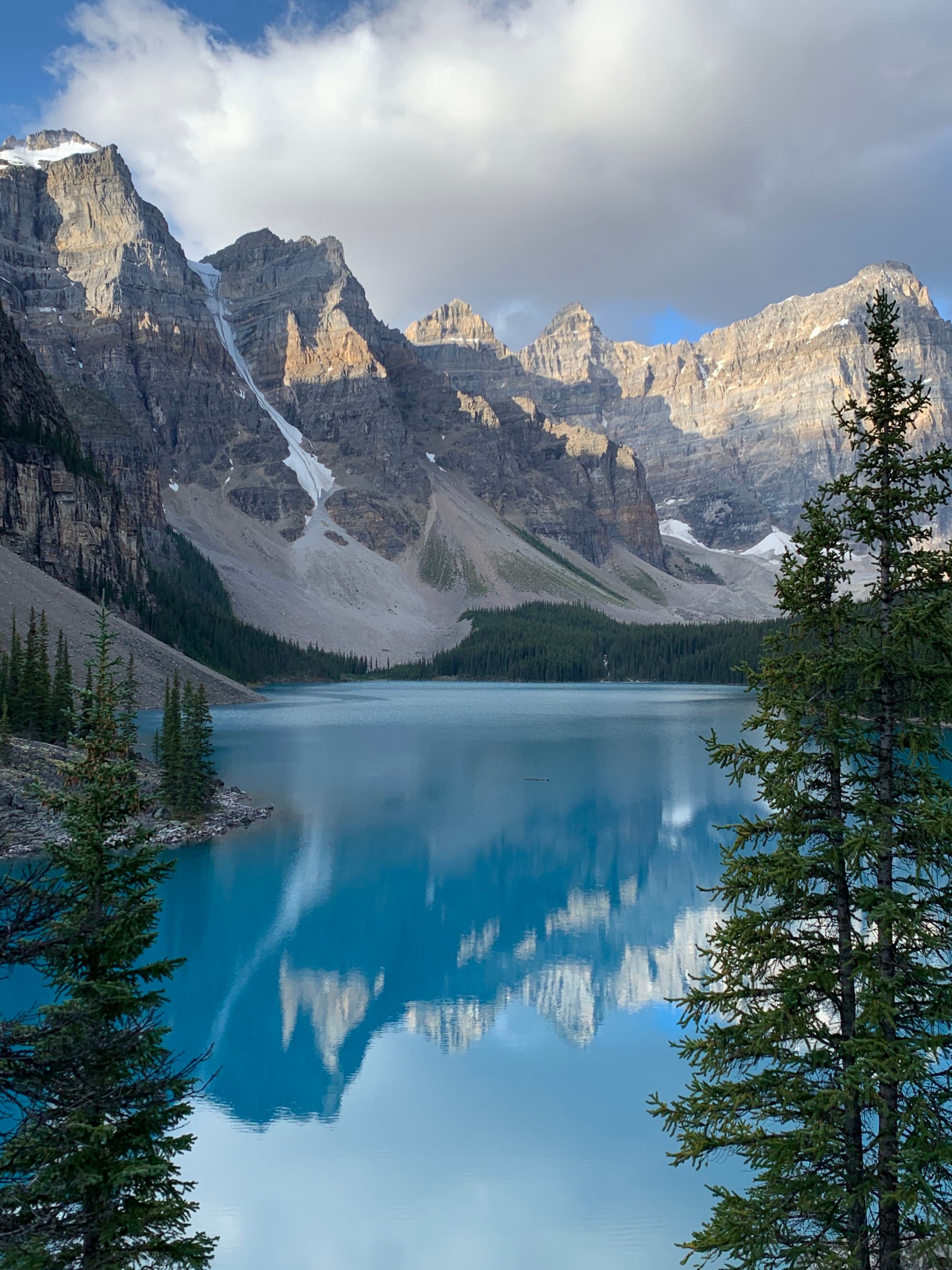 A serene lake reflecting a mountain, framed by evergreen branches, under a partly cloudy sky.