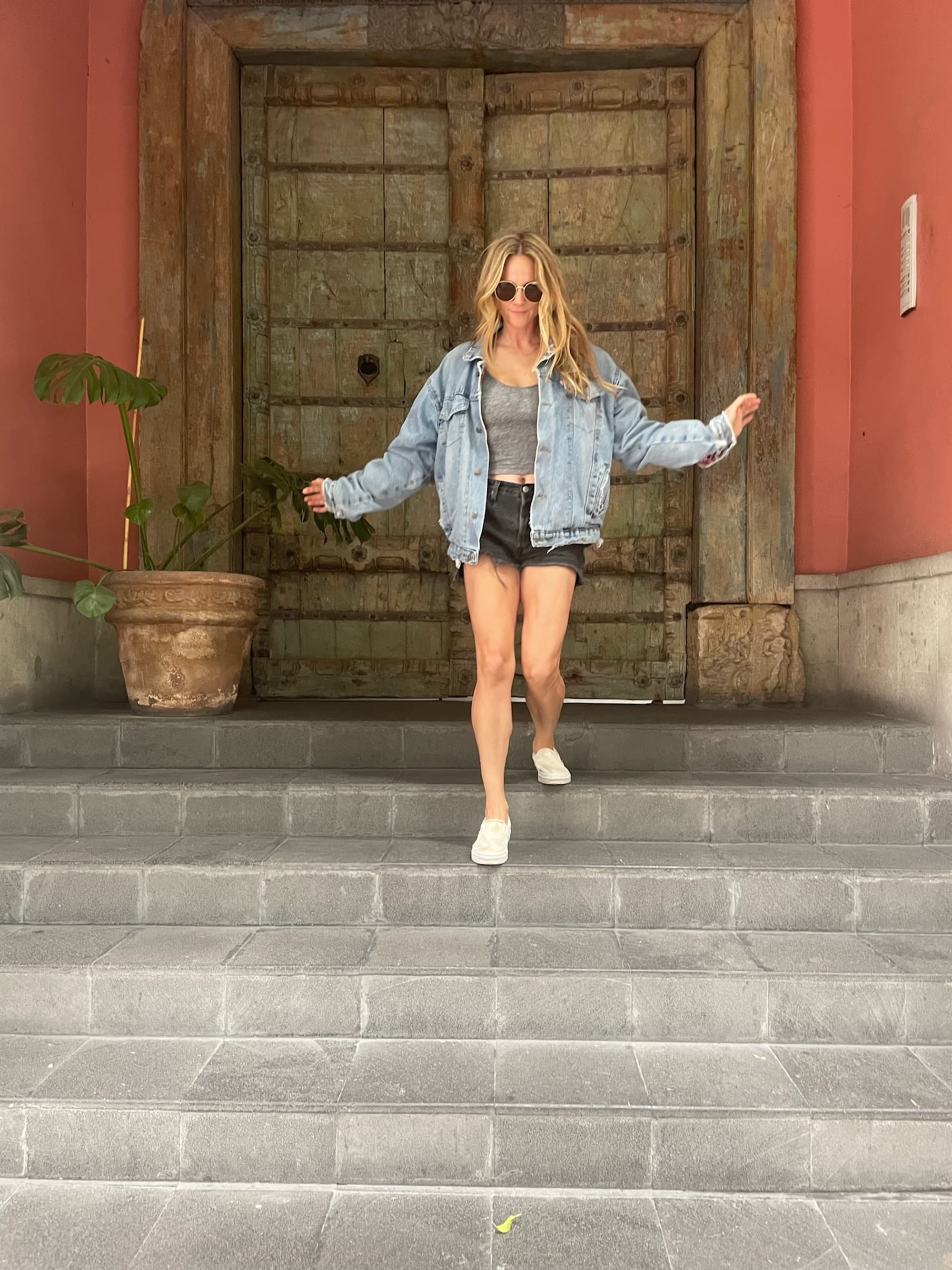 An individual in a denim jacket and black shorts standing on steps in front of a rustic wooden door.