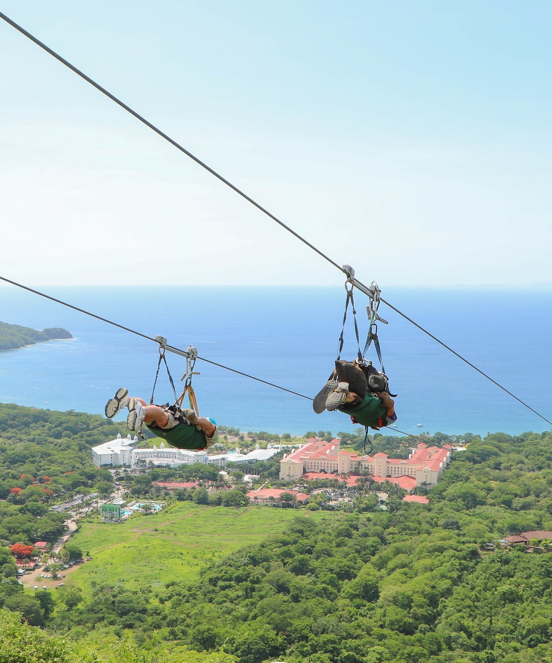 Two people ziplining on a downward slope towards the ocean.