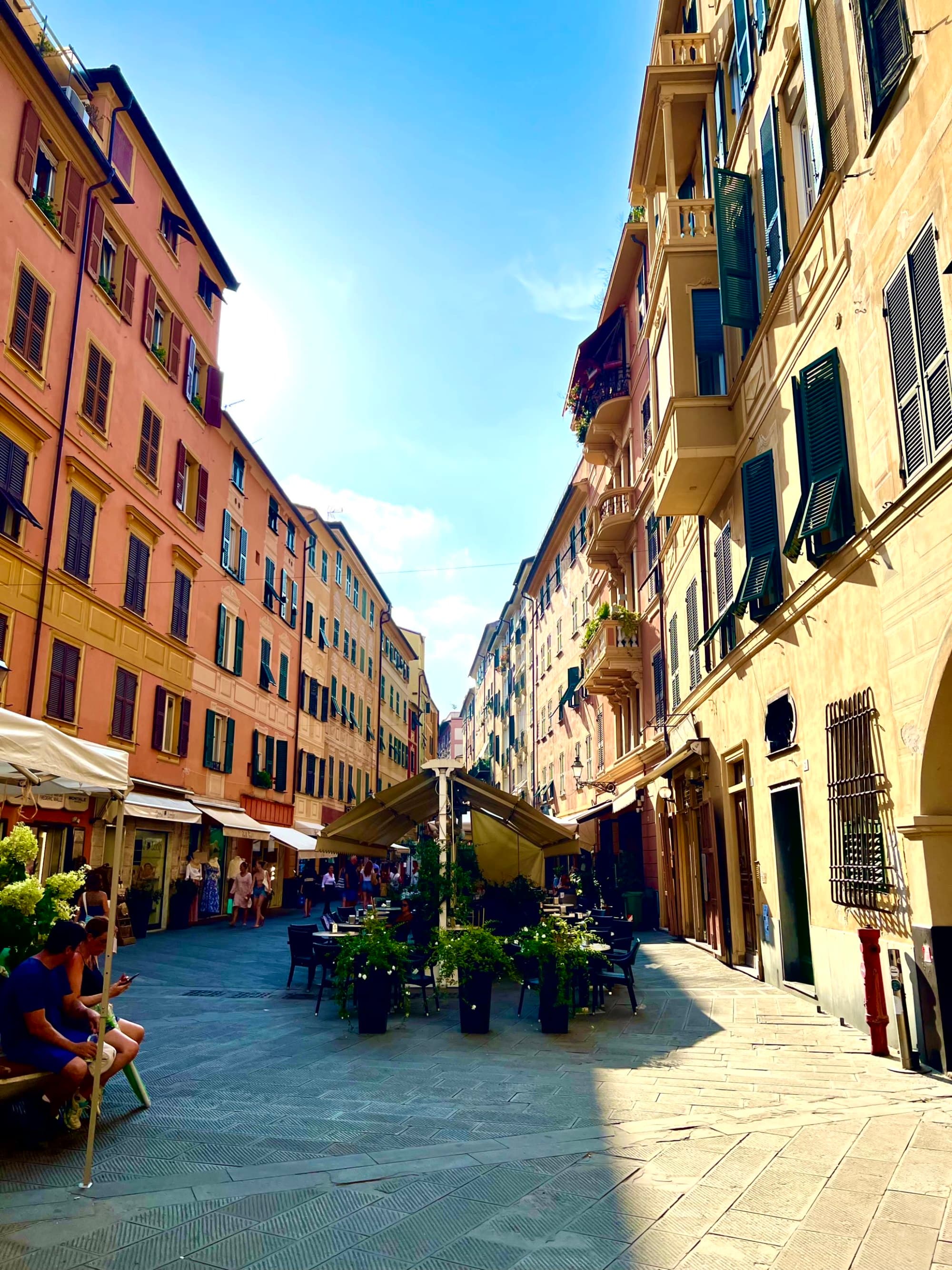 A vibrant street scene with multi-story buildings, outdoor café seating, and a clear blue sky.