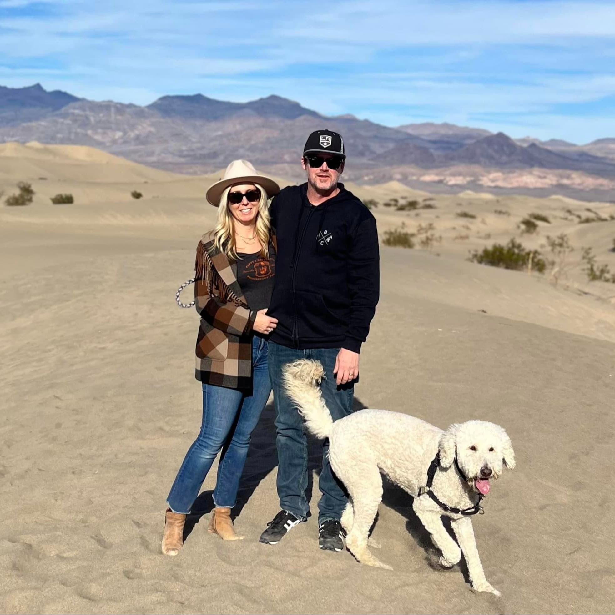 The image shows two individuals with obscured faces and a dog standing in a desert landscape.