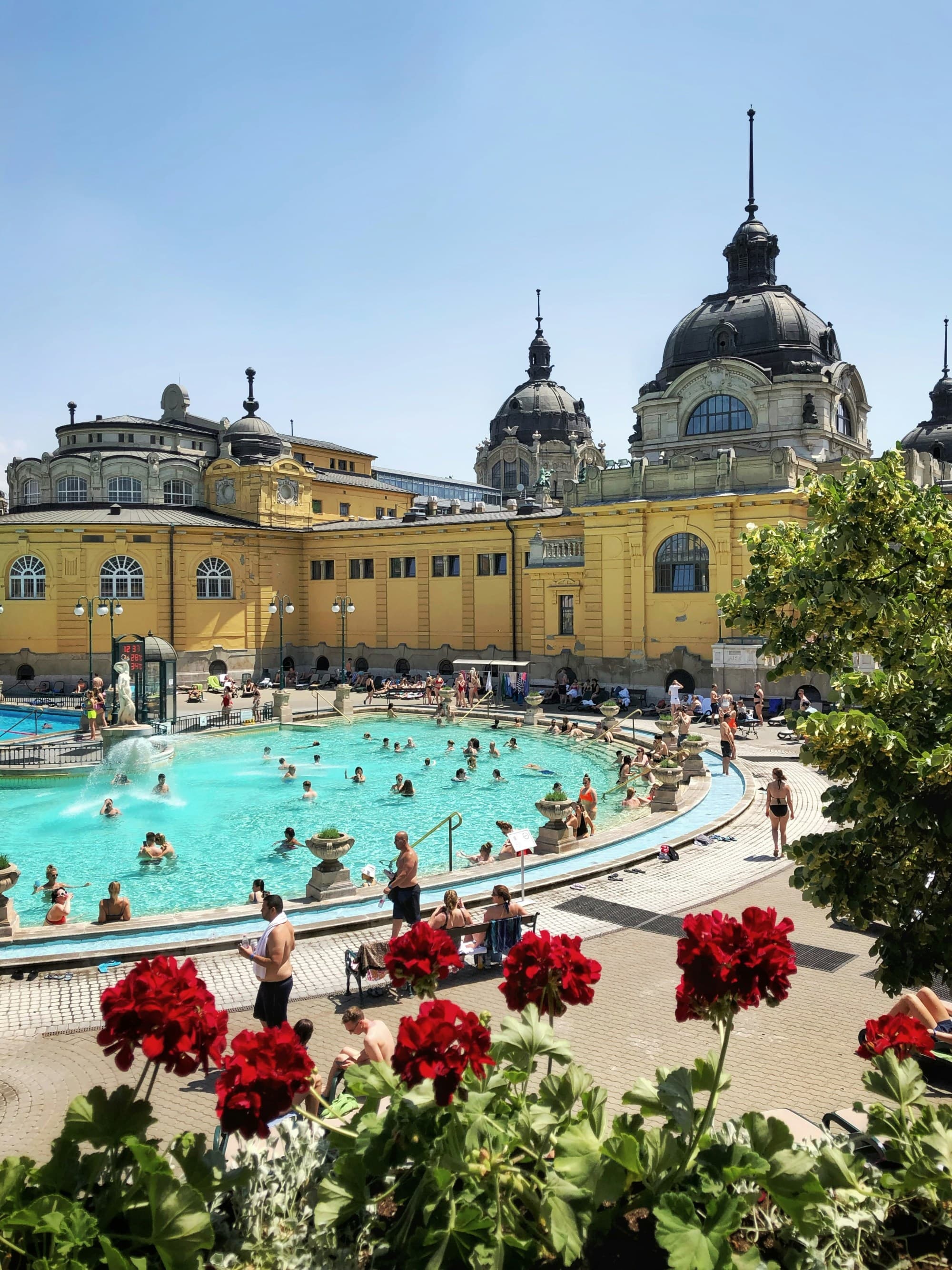 This image shows an outdoor thermal bath with people enjoying the water, set against an ornate, yellow building with domes.