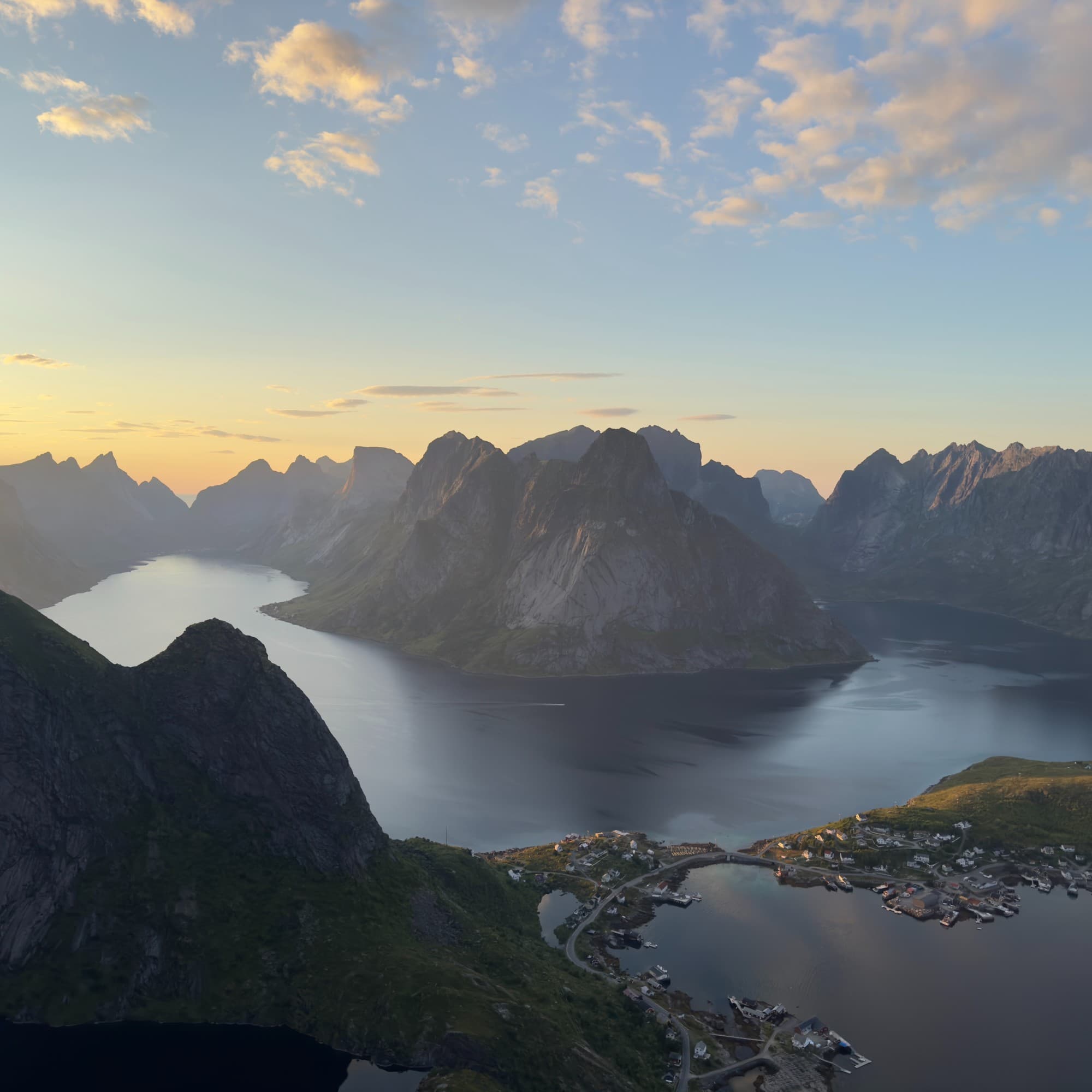 A fjord surrounded by rugged mountains during sunset, with a small settlement at the water’s edge.