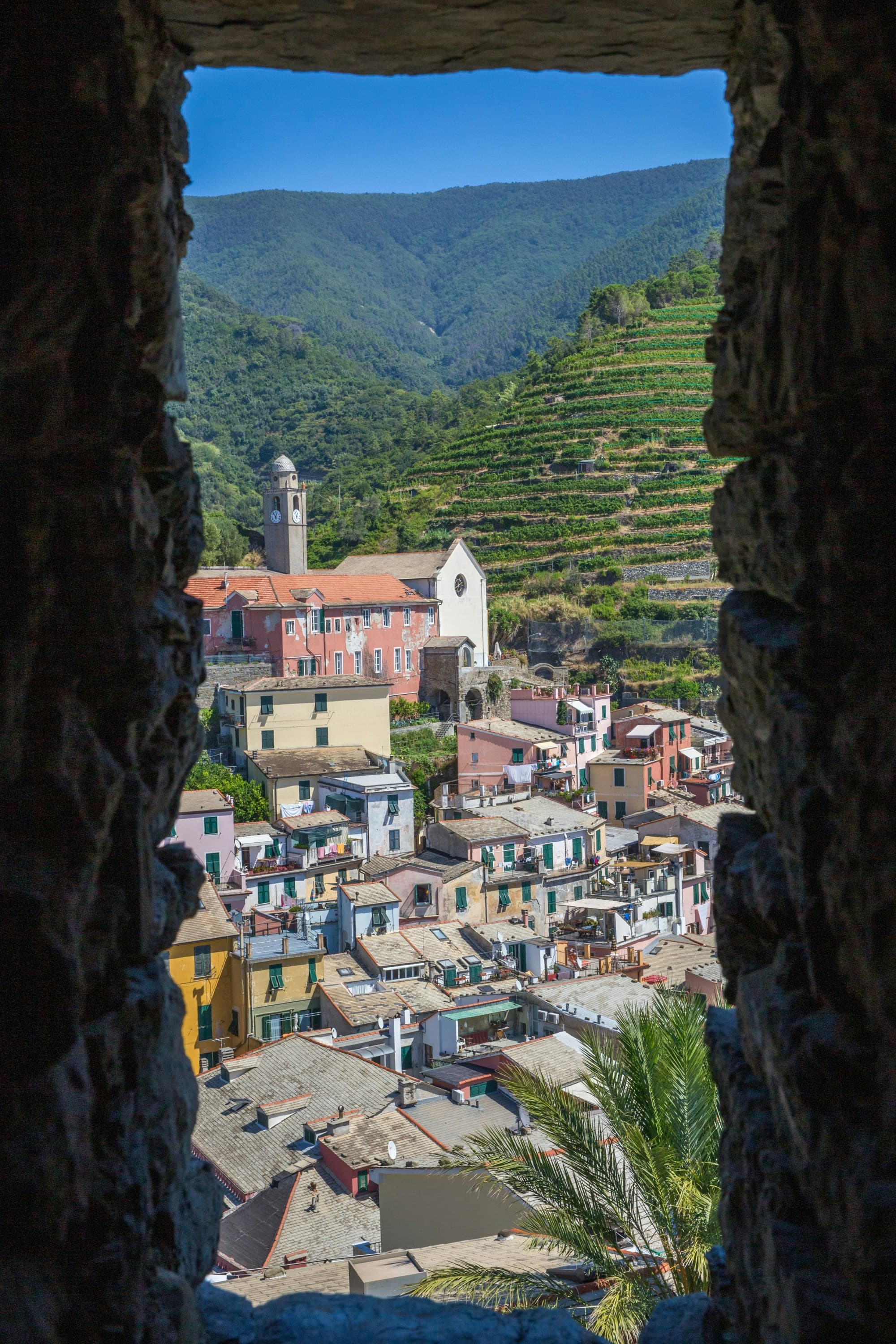 The image shows a scenic view of a colorful village with buildings and a church tower, nestled in a green valley, framed by the silhouette of an arched stone window.