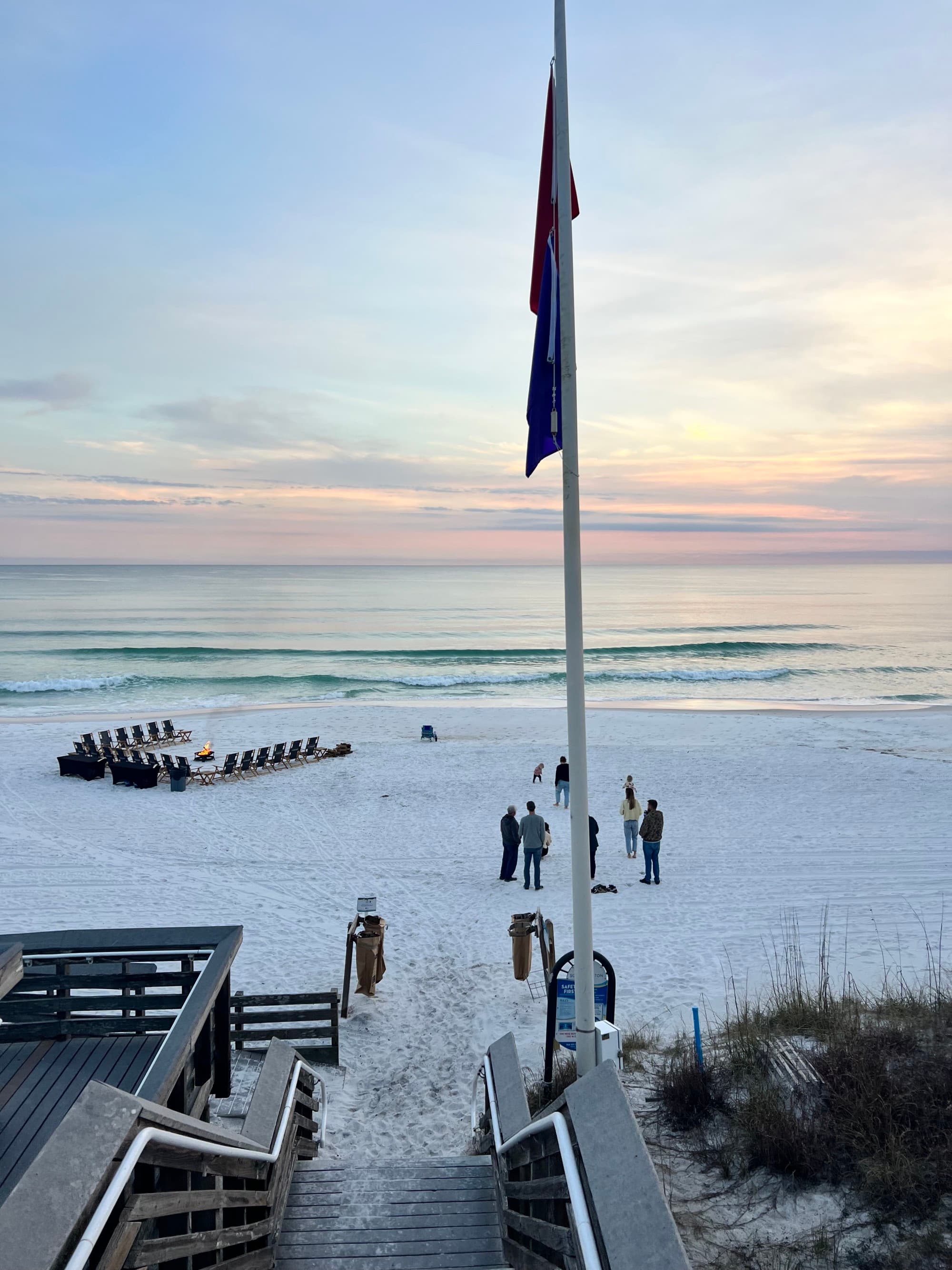 The image shows a serene white sand beach scene with people, a wooden boardwalk, a flag and chairs arranged in rows, set against a beautiful sunset or sunrise.