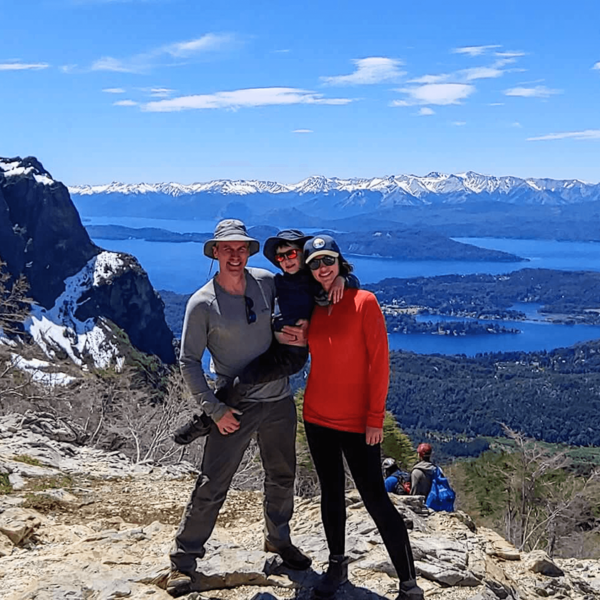 Two individuals are standing on a mountain trail with a panoramic view of a lake and mountains in the background.