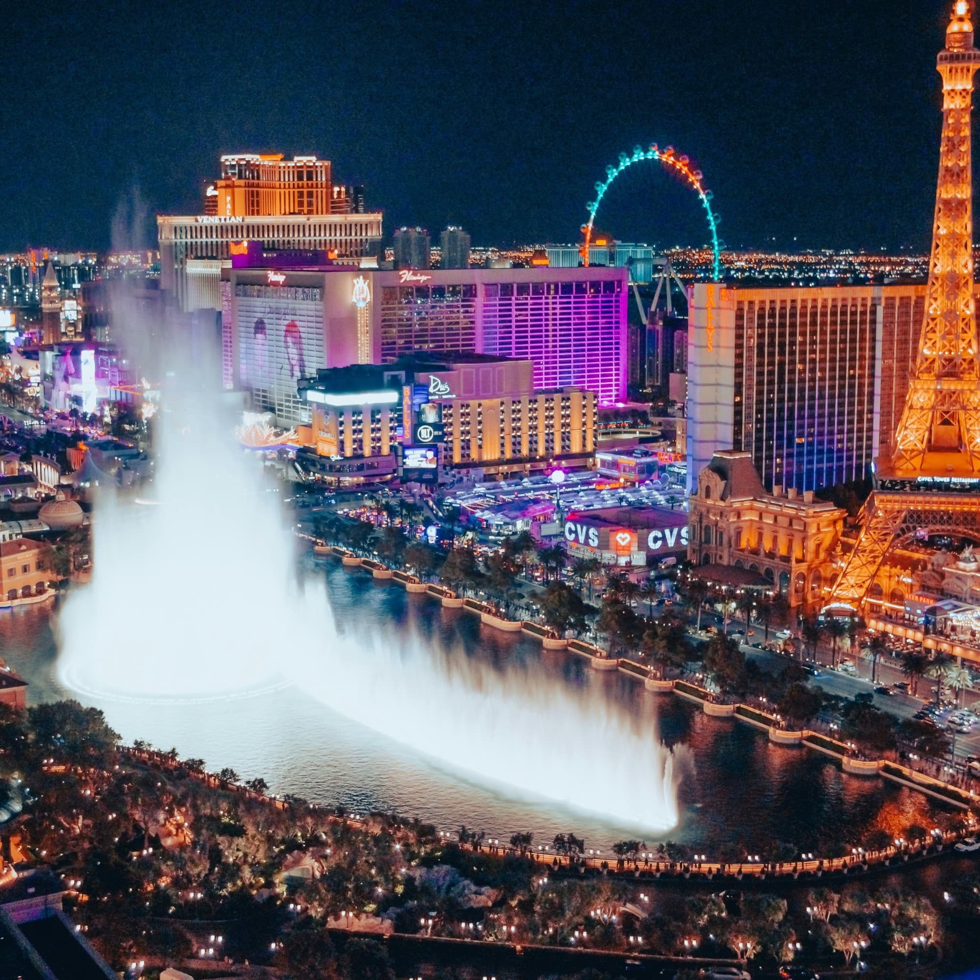 The image shows a vibrant nighttime aerial view of the Las Vegas Strip, with illuminated buildings and a large fountain show.