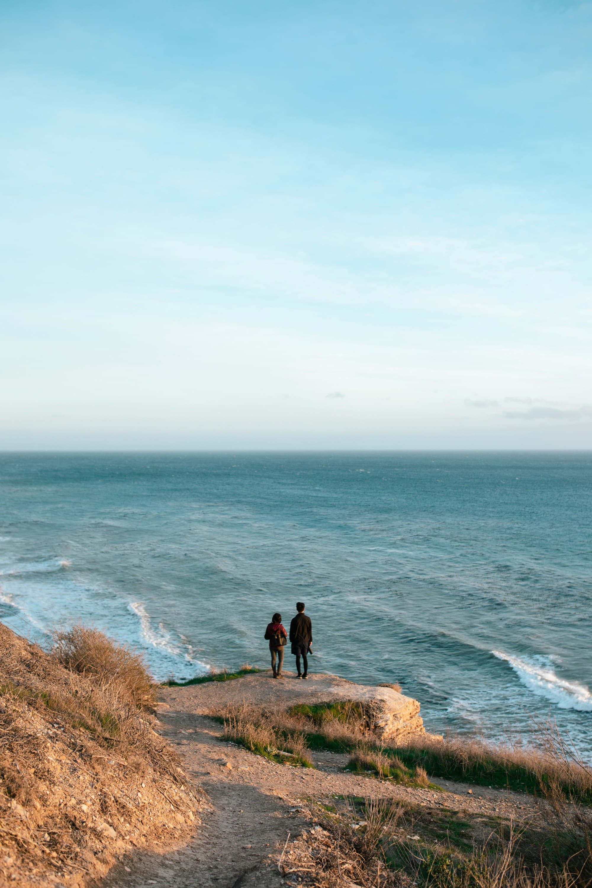 two people standing on a cliffside next to the ocean during the daytime