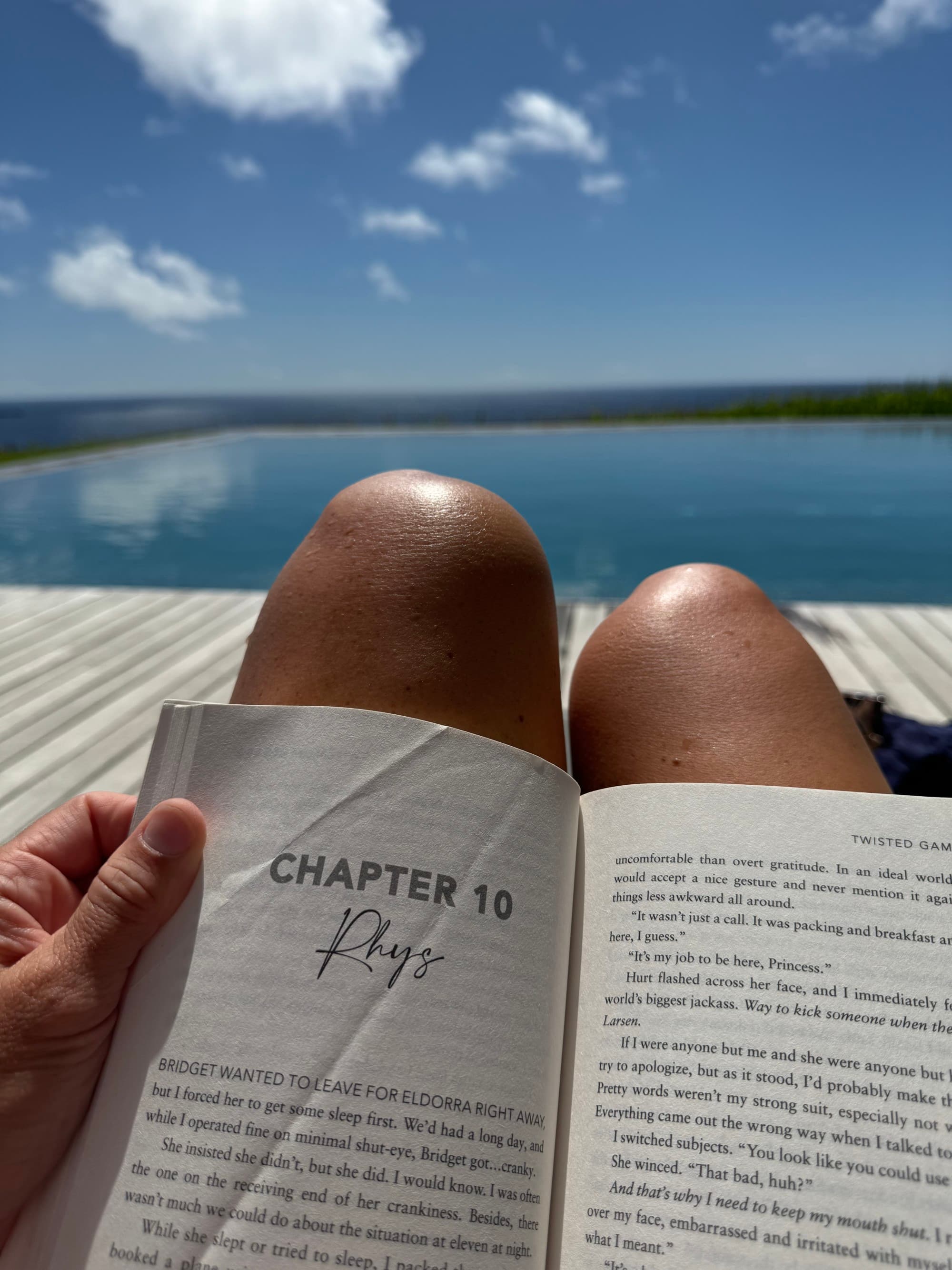 A person’s legs with a book on top are seen against the backdrop of a tranquil blue sea and sky.