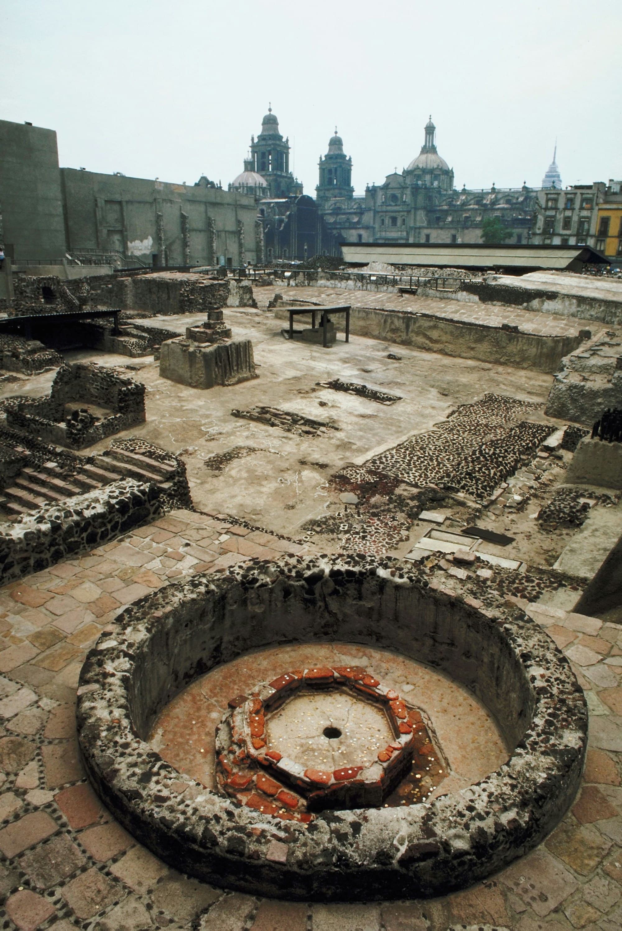 An archaeological site with ancient ruins in the foreground and a modern cityscape with buildings and a church in the background.