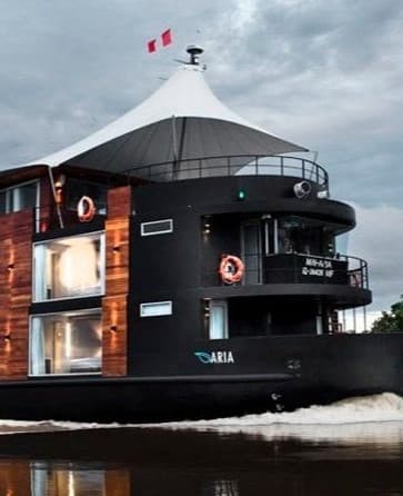 A large, multi-story riverboat with a sleek black exterior and wooden accents cruises on water under a cloudy sky.