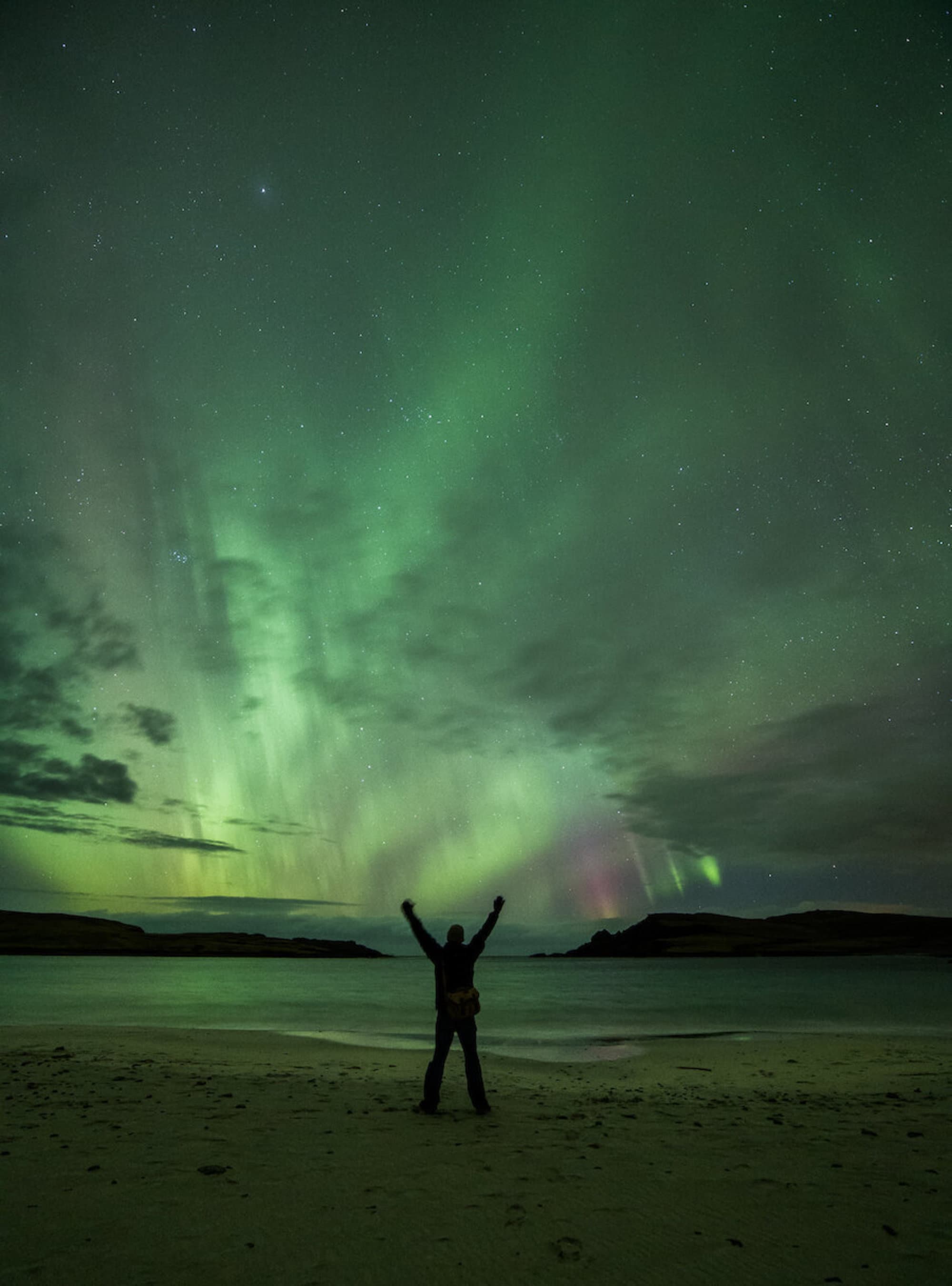 The image shows a person with arms raised standing on a beach at night, under a sky illuminated by the aurora borealis.