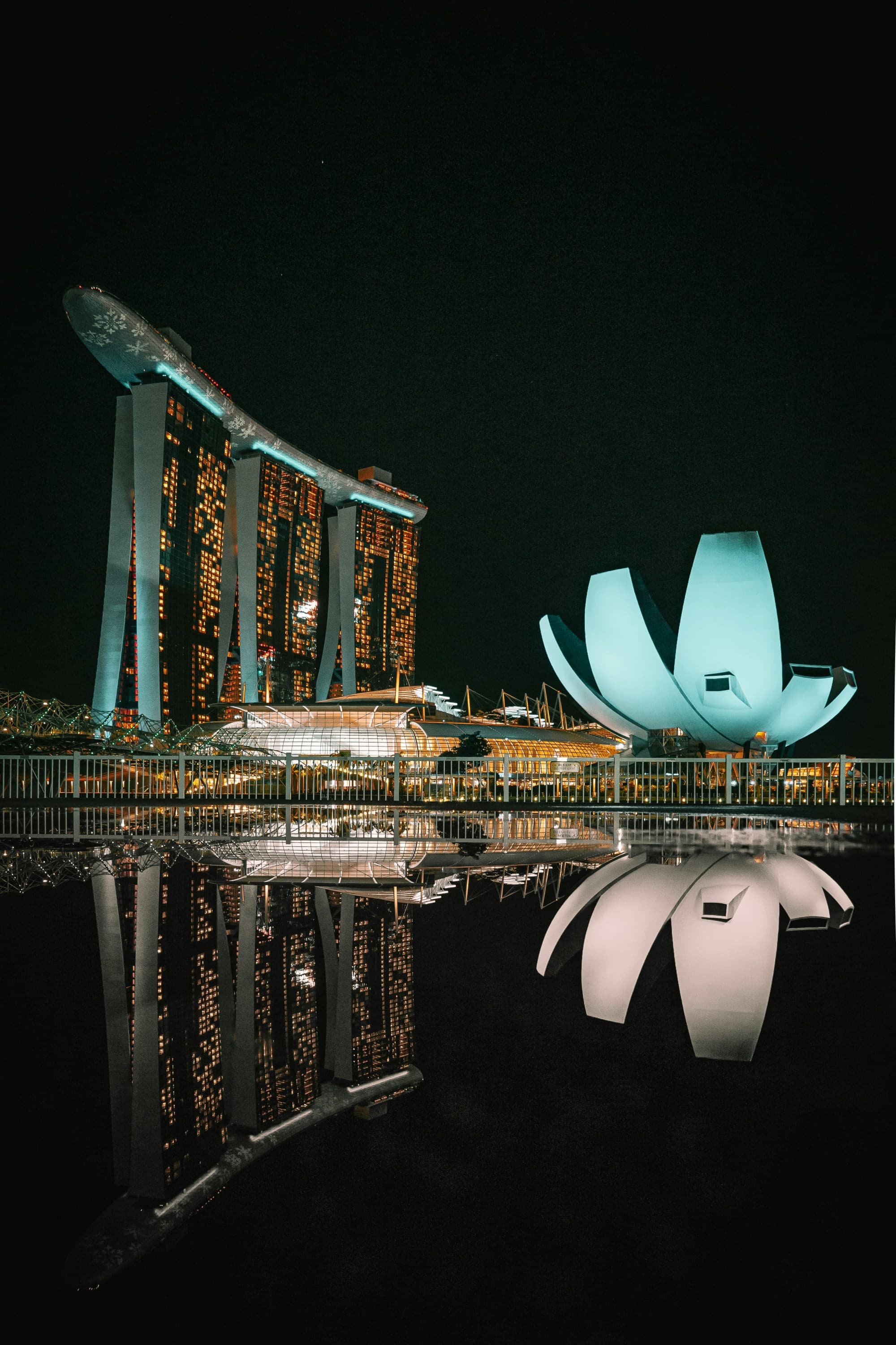 The image shows the Marina Bay Sands Hotel and ArtScience Museum in Singapore at night, with their reflections visible on the water’s surface.