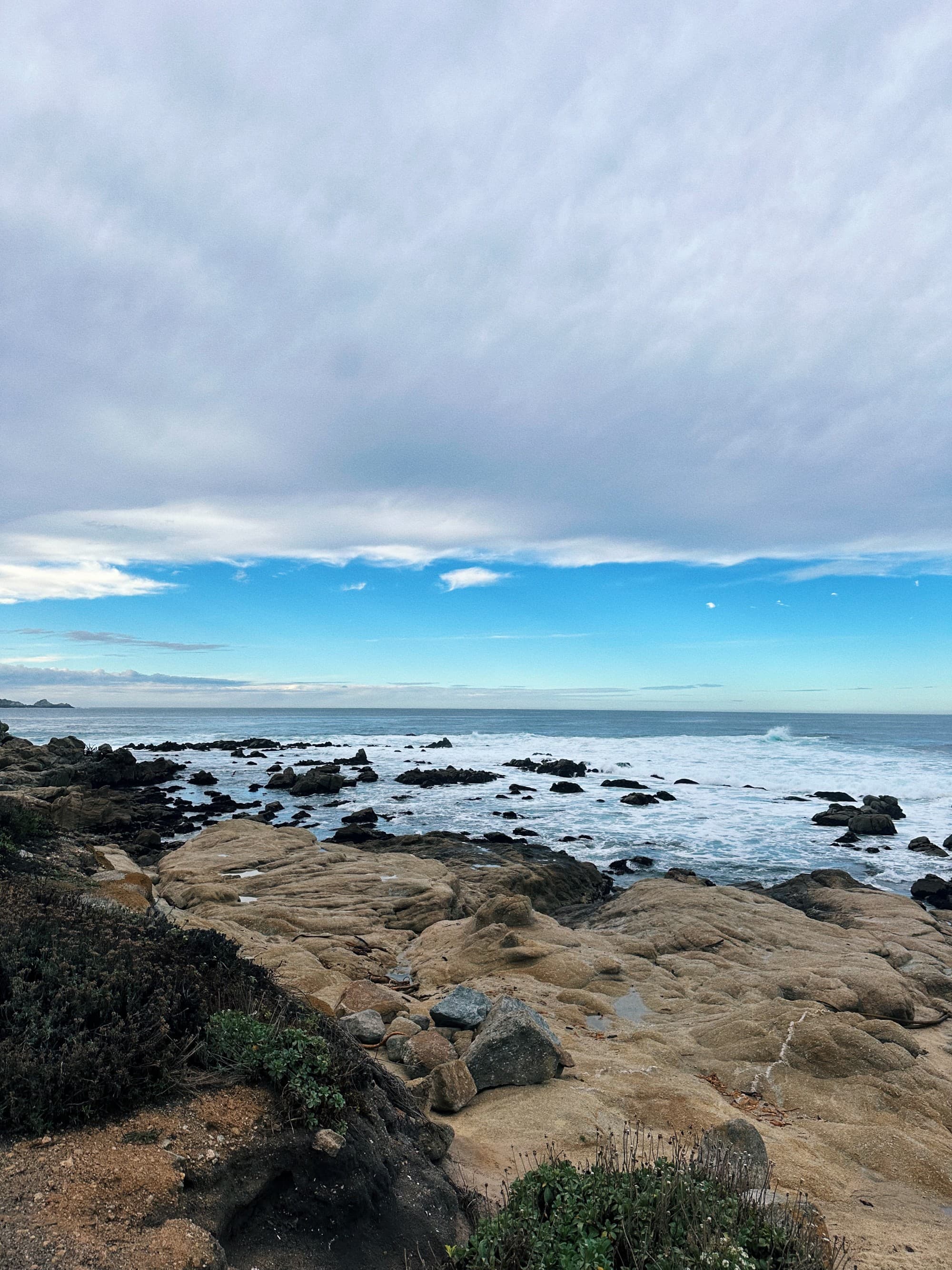 The image depicts a coastal scene with rocky shores, ocean waves and a cloudy blue sky.