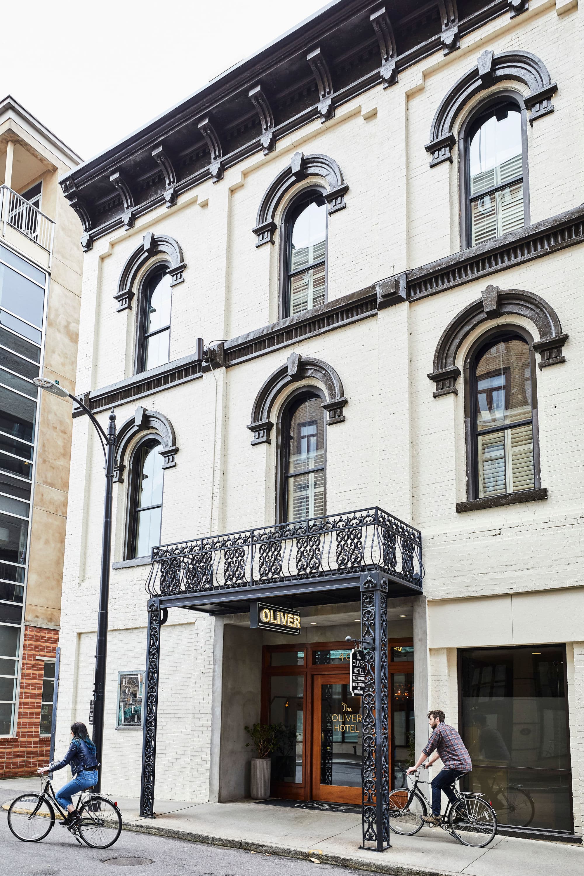 The entrance of a three story white building with black trim called the Oliver hotel and two people riding bikes during the day.