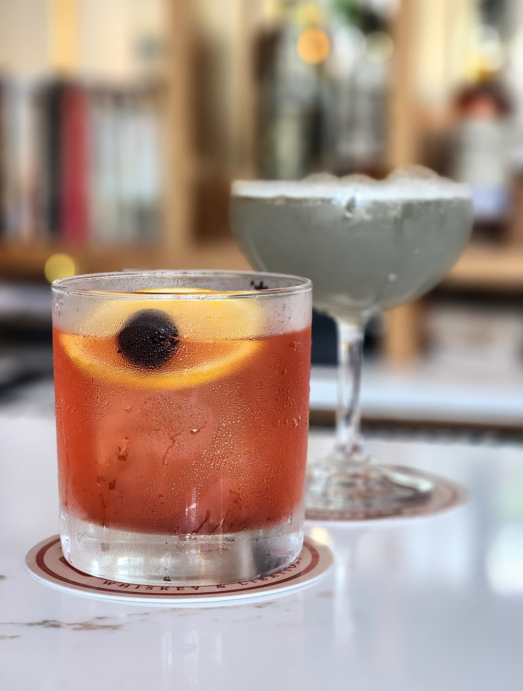 The image shows a chilled beverage with ice, garnished with an orange slice and a dark-colored berry, set on a coaster.