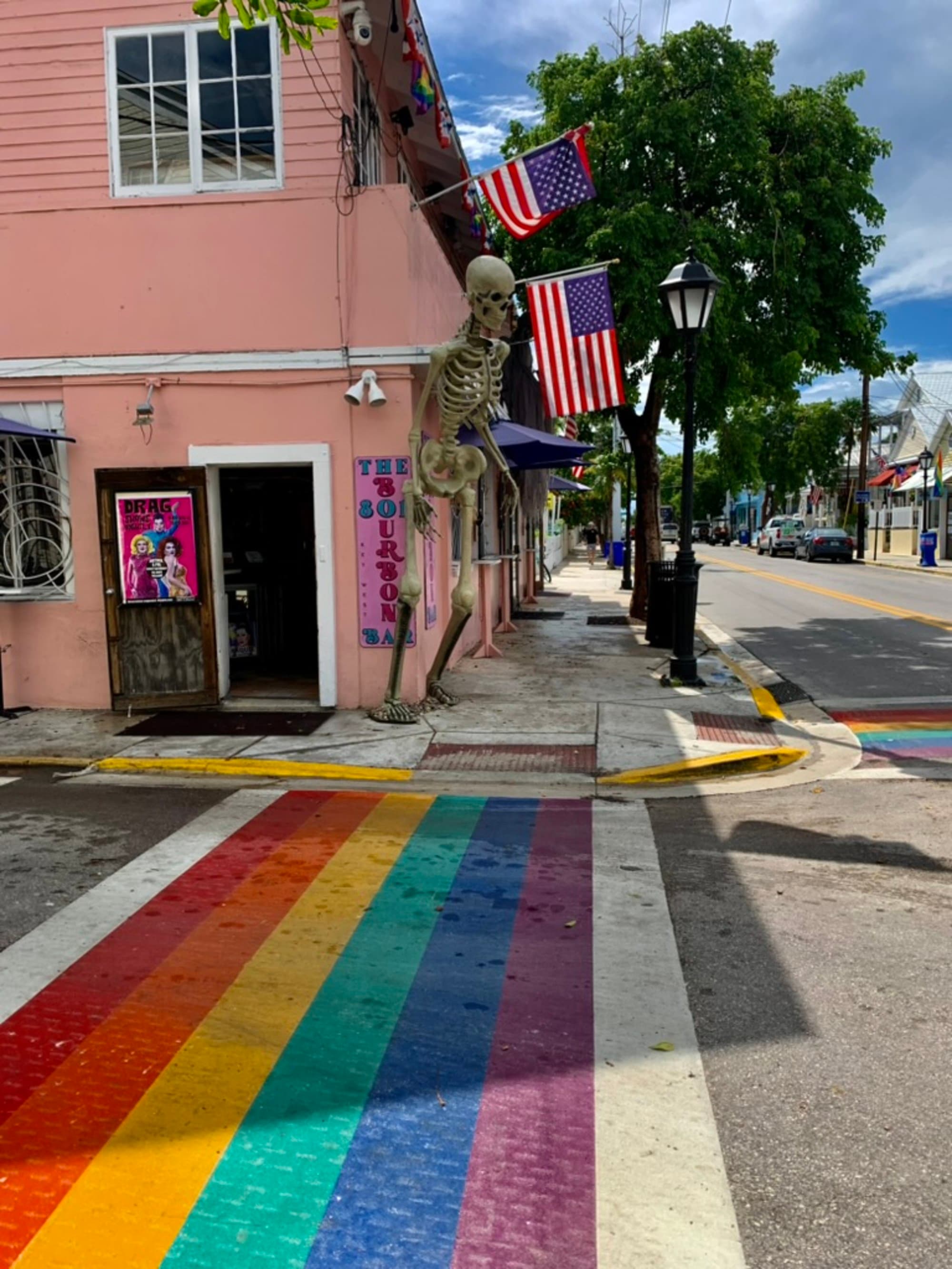 The image shows a vibrant street corner with a rainbow-colored crosswalk, a pink building adorned with American flags, a skeleton decoration, and a lamppost.