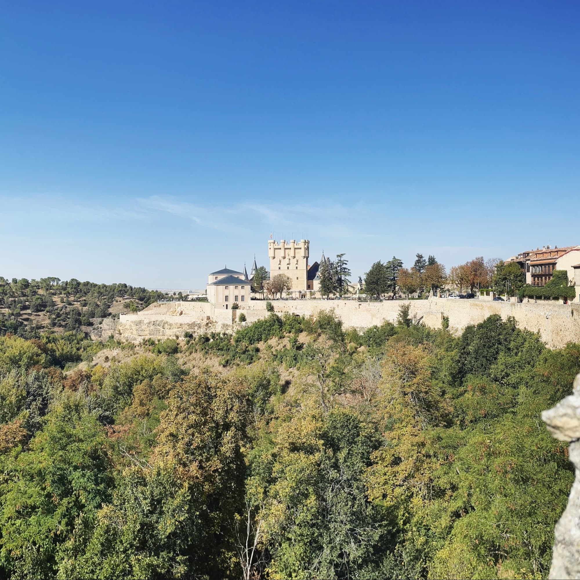 A historic stone building with towers is surrounded by lush greenery under a clear blue sky.