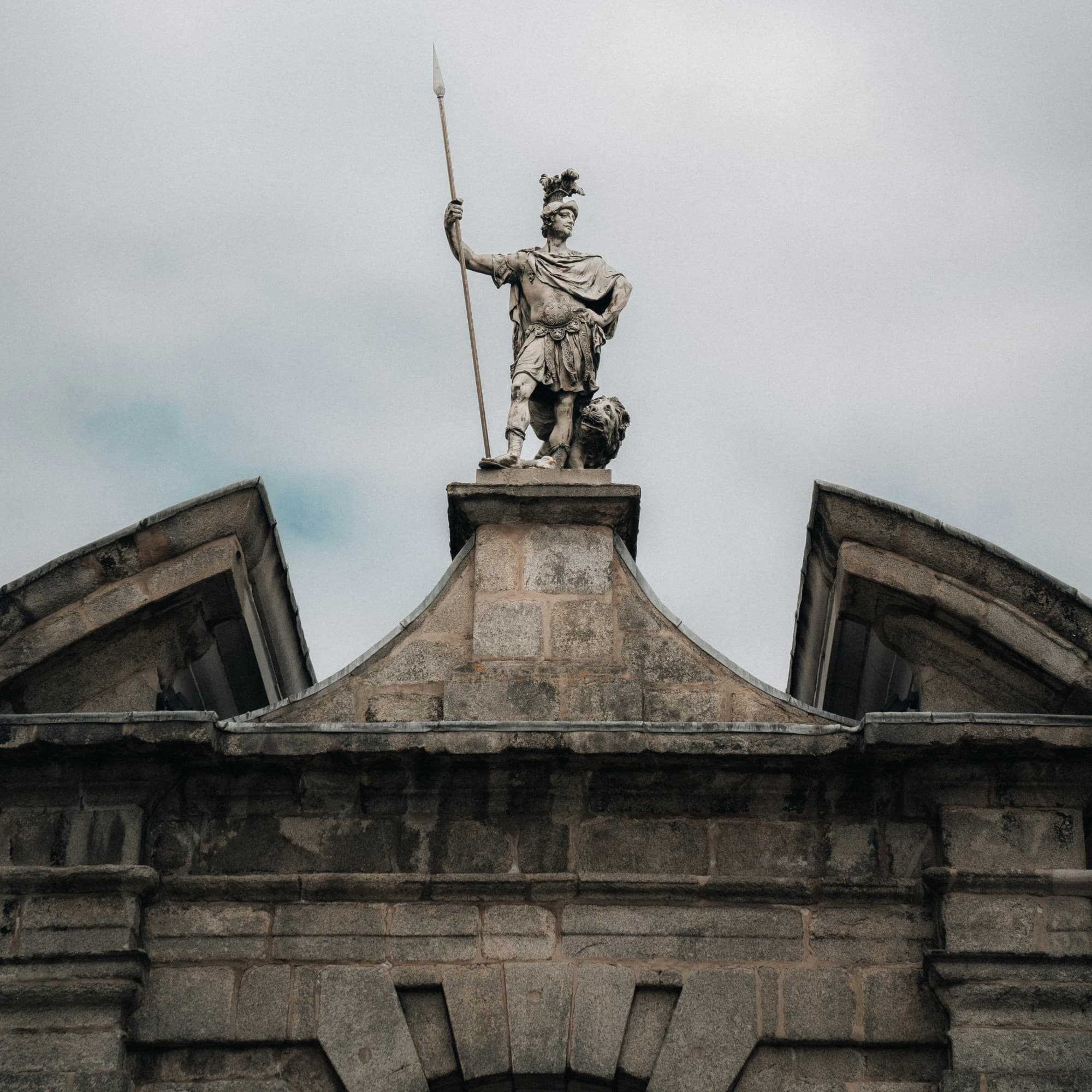 The image displays a statue of a warrior with a spear atop an ornate stone pediment against a cloudy sky.