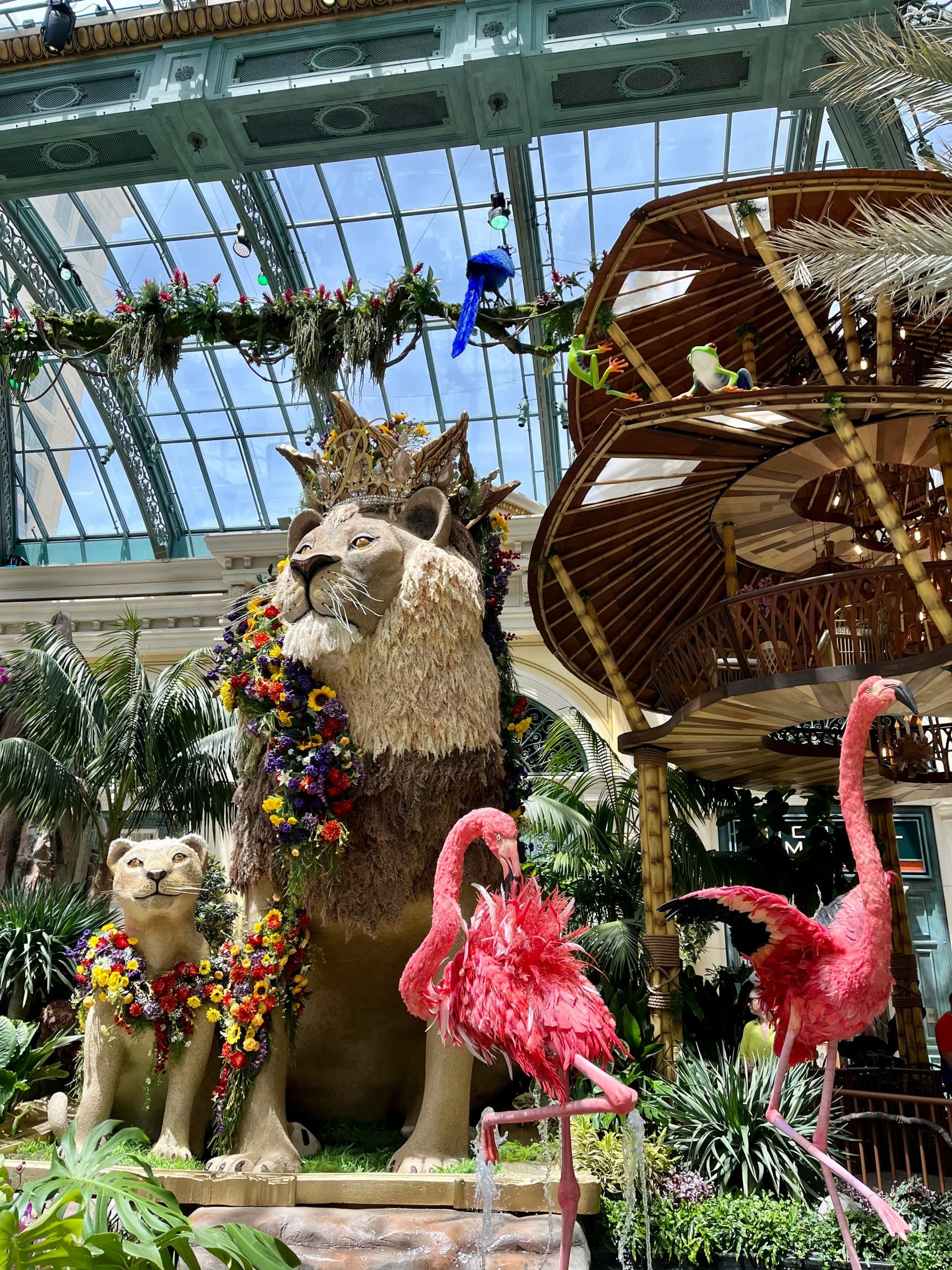 A vibrant indoor garden features a large lion statue adorned with colorful flowers, surrounded by smaller animal figures and lush greenery under a glass ceiling.