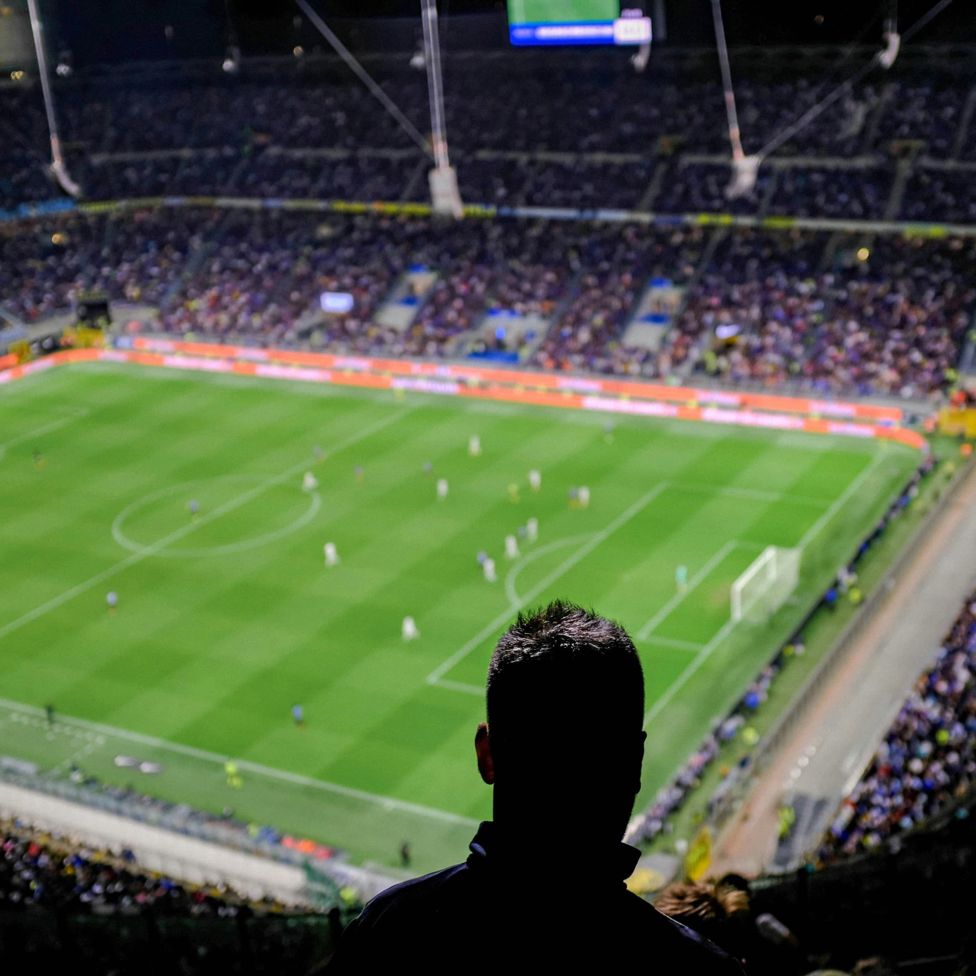 An elevated view of a nighttime football match in a packed stadium, with the back of a person’s head in the foreground.