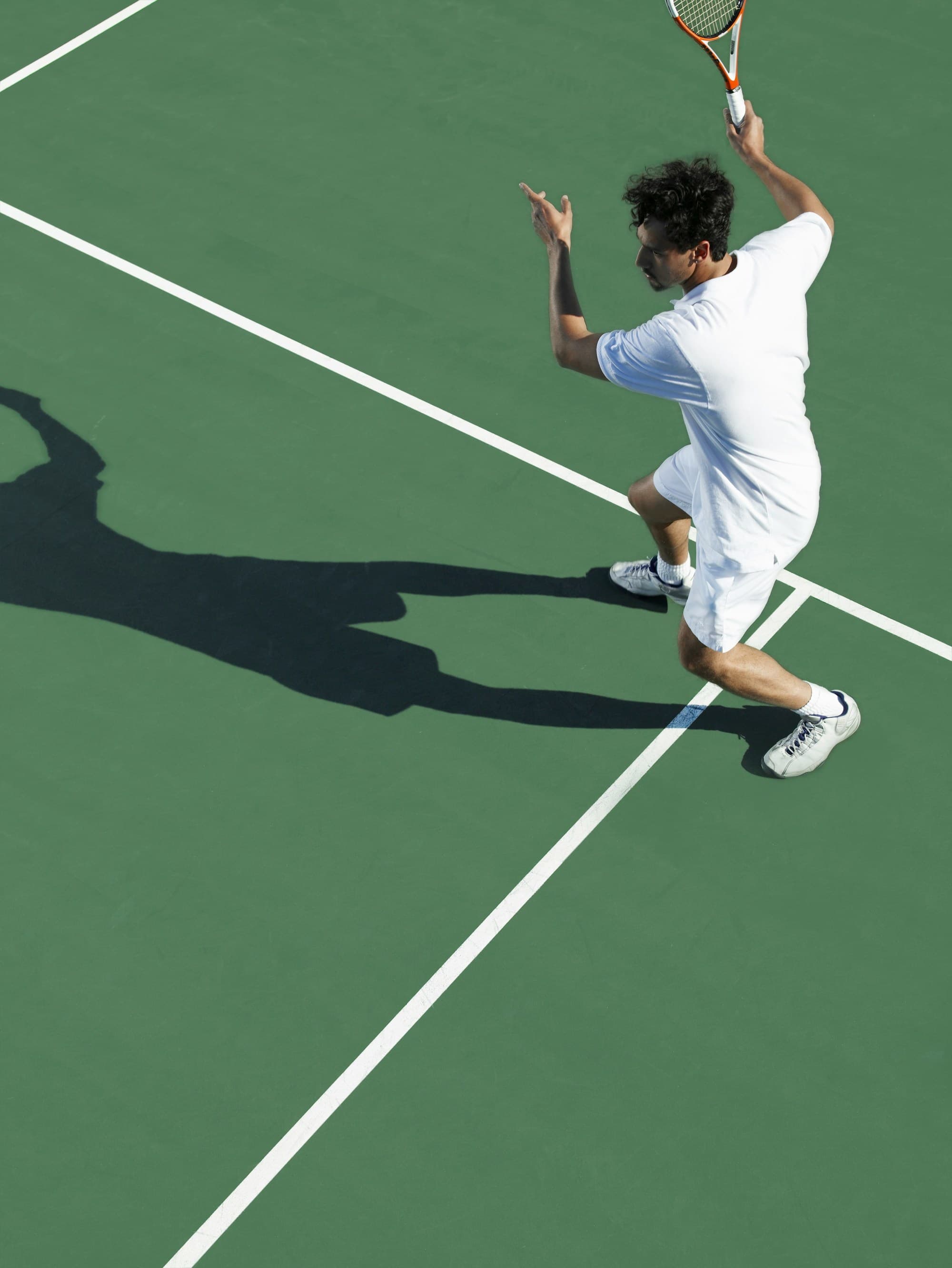 A tennis player in white attire mid-action on a green tennis court on a sunny day.