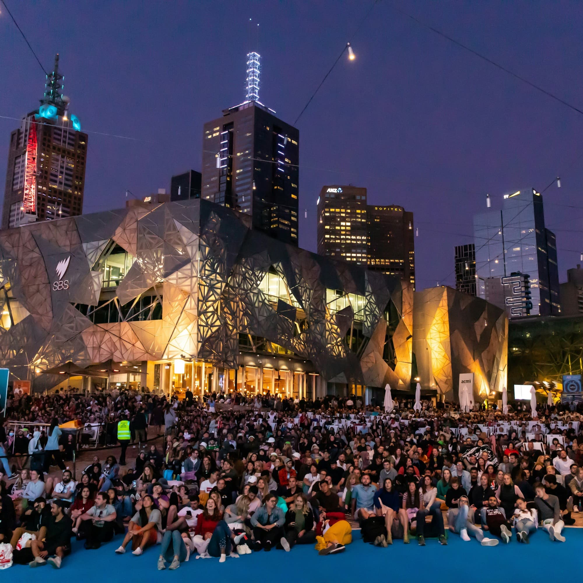 A large crowd sits on the ground at an outdoor event during dusk, with a distinctive, angular building and a city skyline in the background.