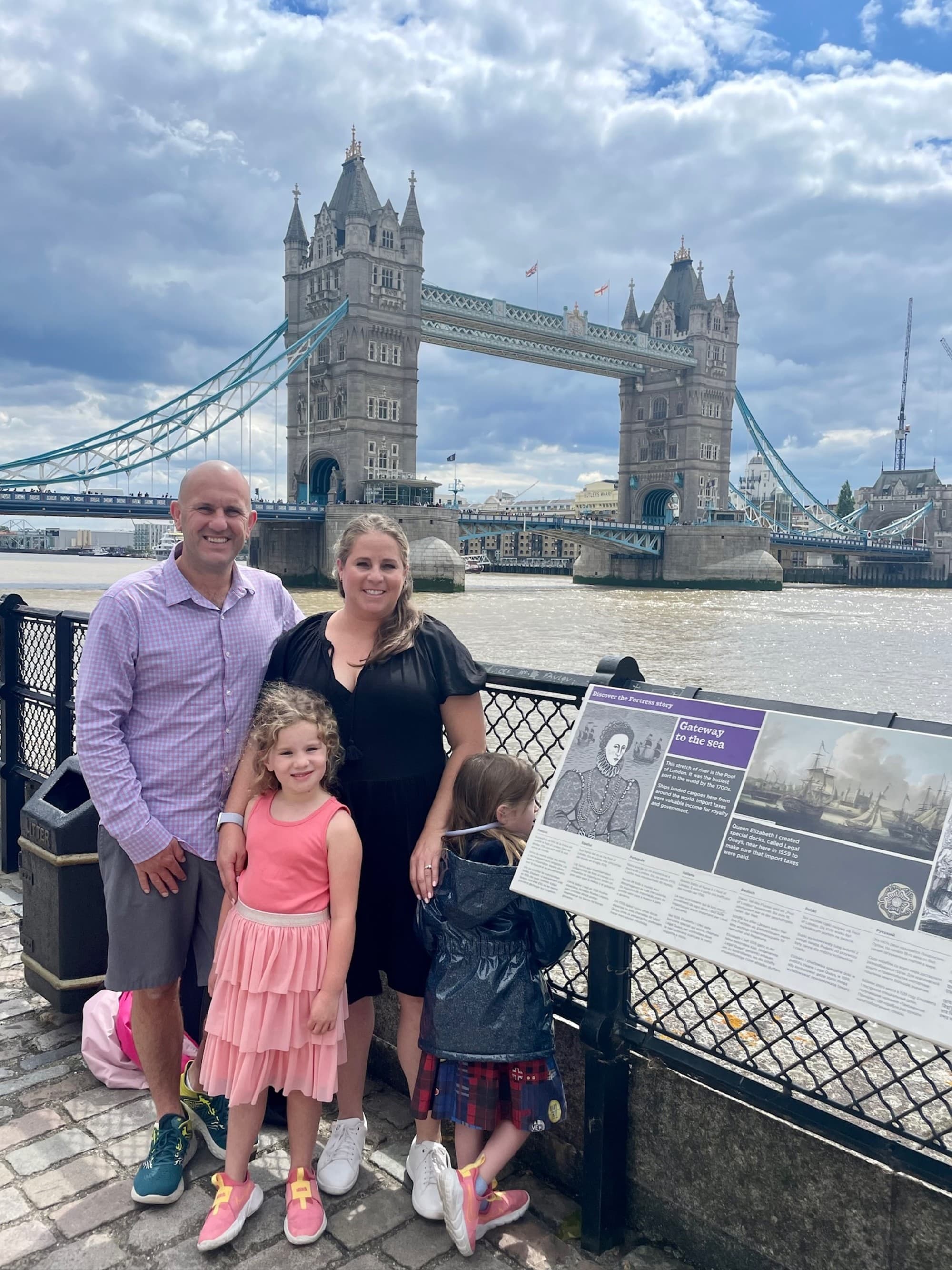 A family posing for a photo in front of the Tower Bridge in London, with an informational plaque in the foreground.