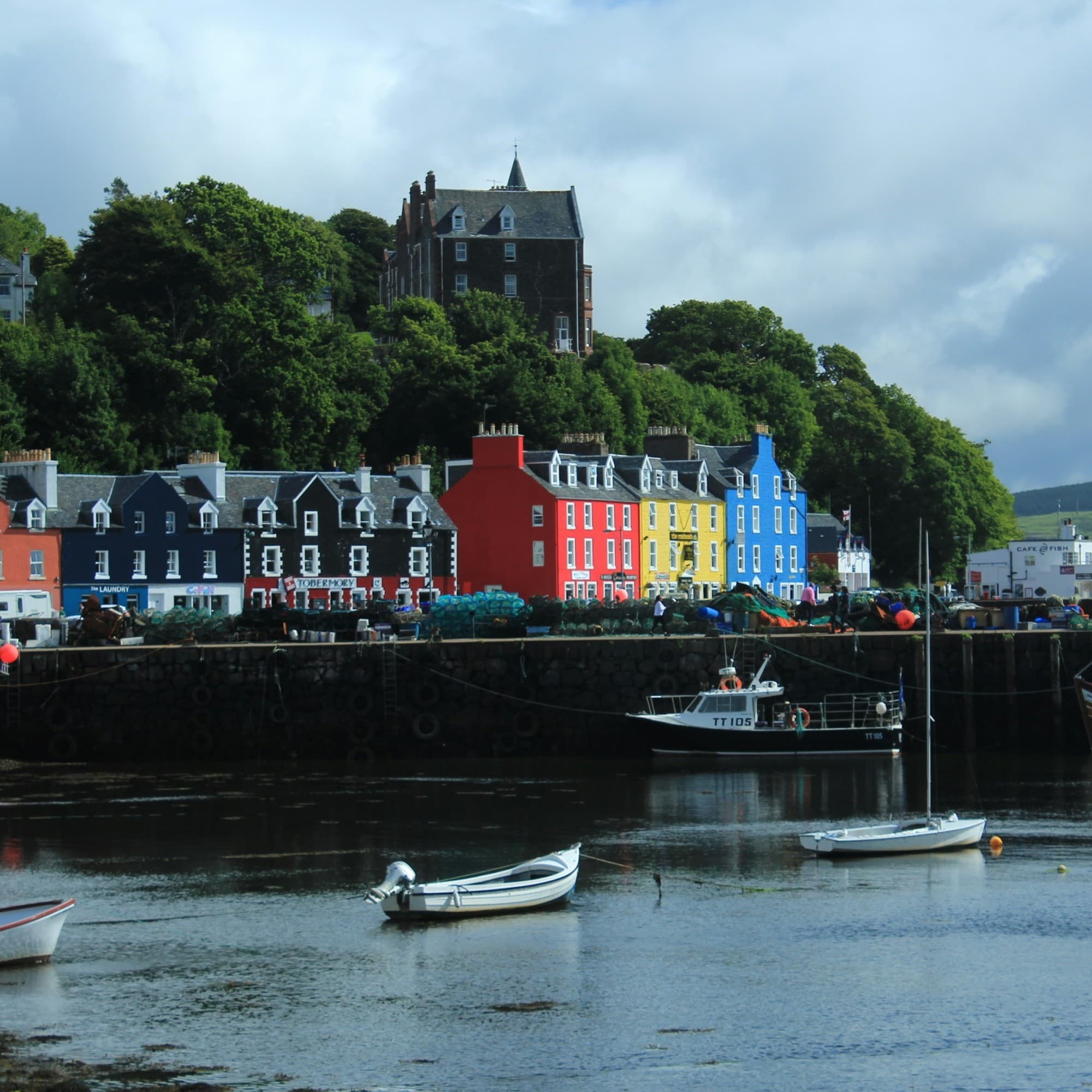 The image shows a scenic waterfront with colorful buildings, boats and a hill with greenery under a cloudy sky.