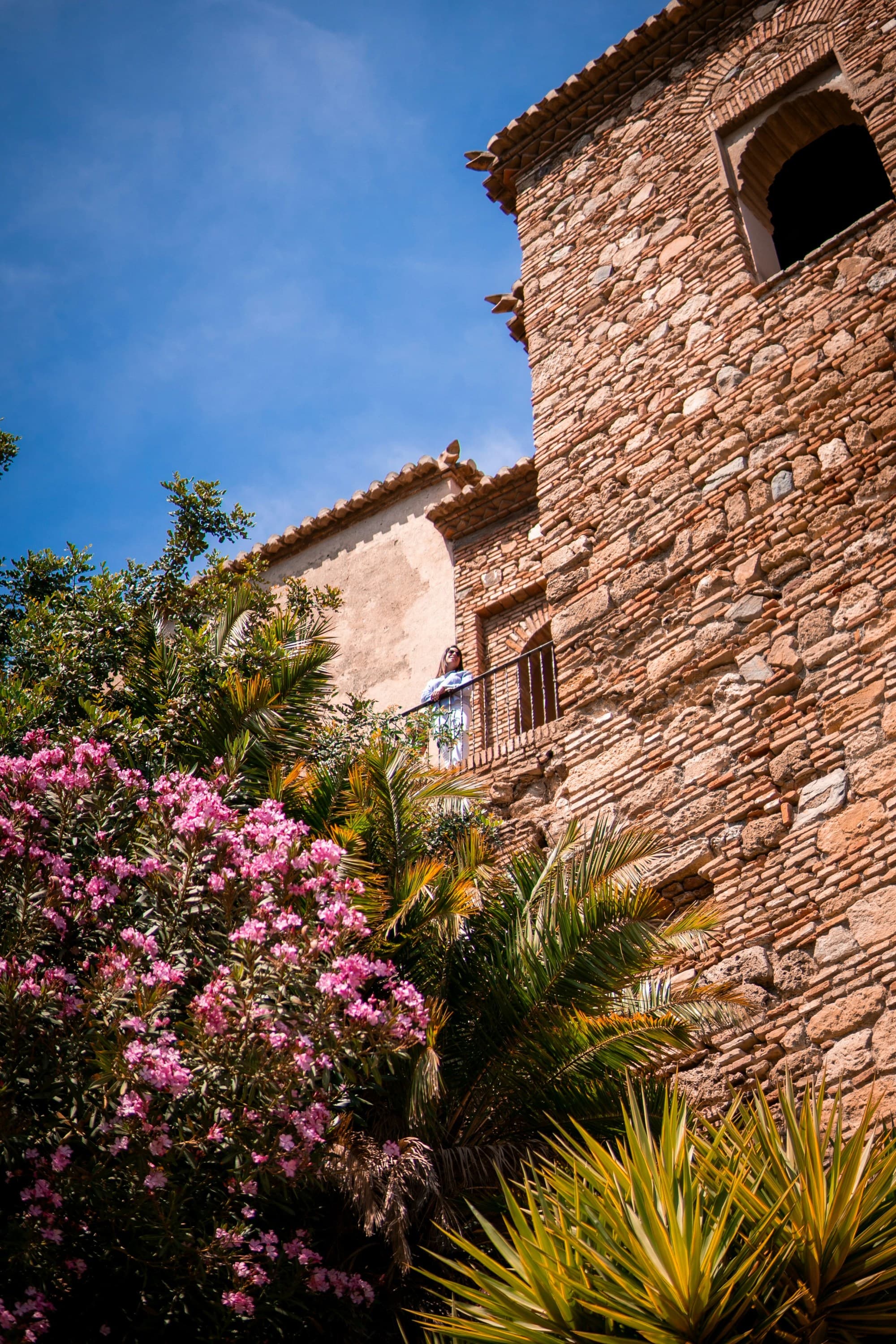 A stone building with a balcony is surrounded by lush greenery and pink flowers under a clear, blue sky.