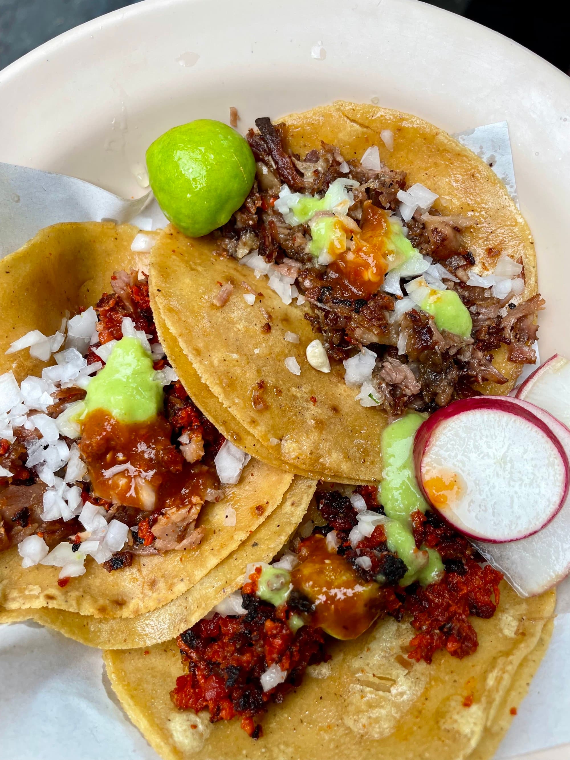 Three tacos with various toppings on a white plate, accompanied by a slice of lime and radish.