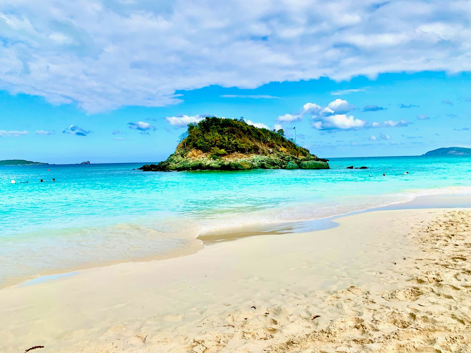 An island dotted with lush foliage sits surrounded by lapping waves just off the beach shore on a sunny day.