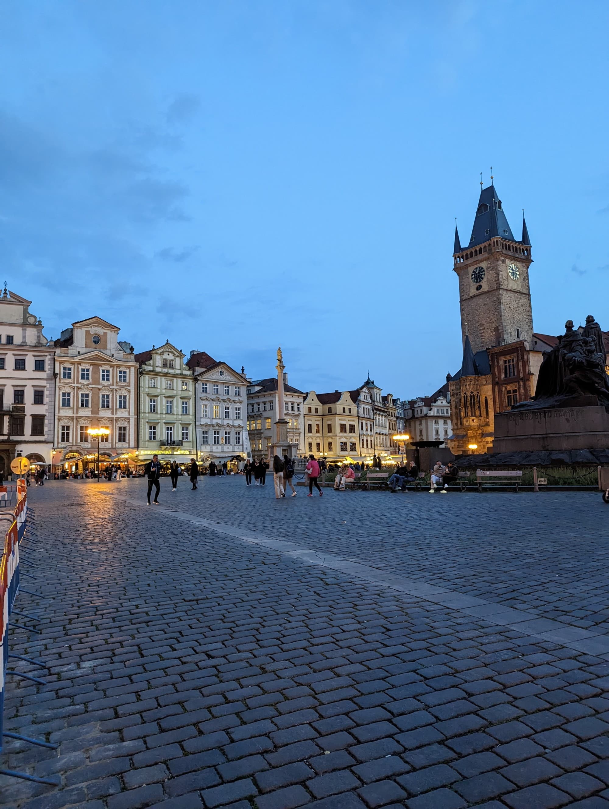 A view of a cobblestone street, old clock tower and stone buildings in the evening lit up with street lamps near people walking around.