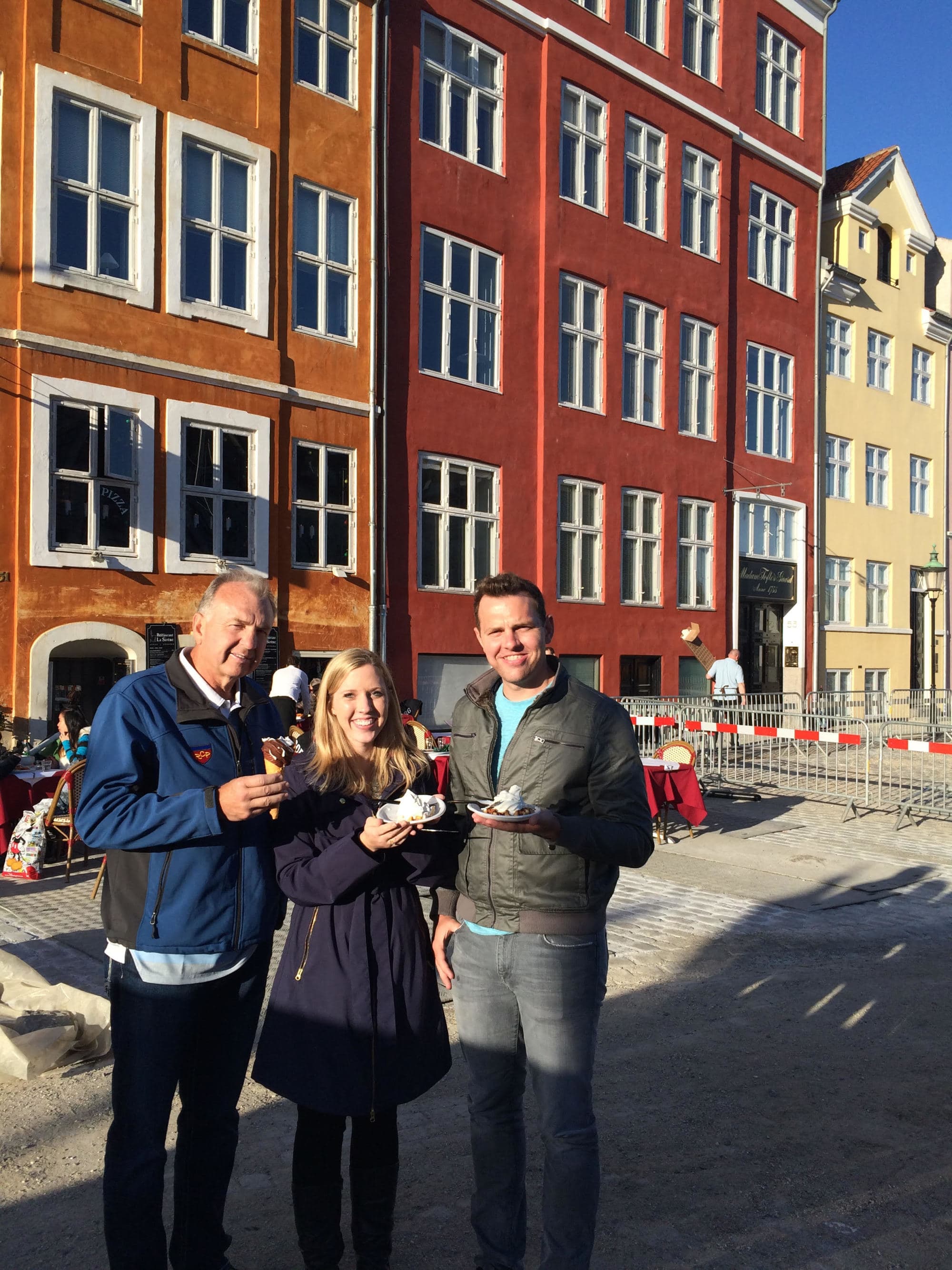 Two men and a woman standing in front of a brown building with food in hand on a street during daytime.