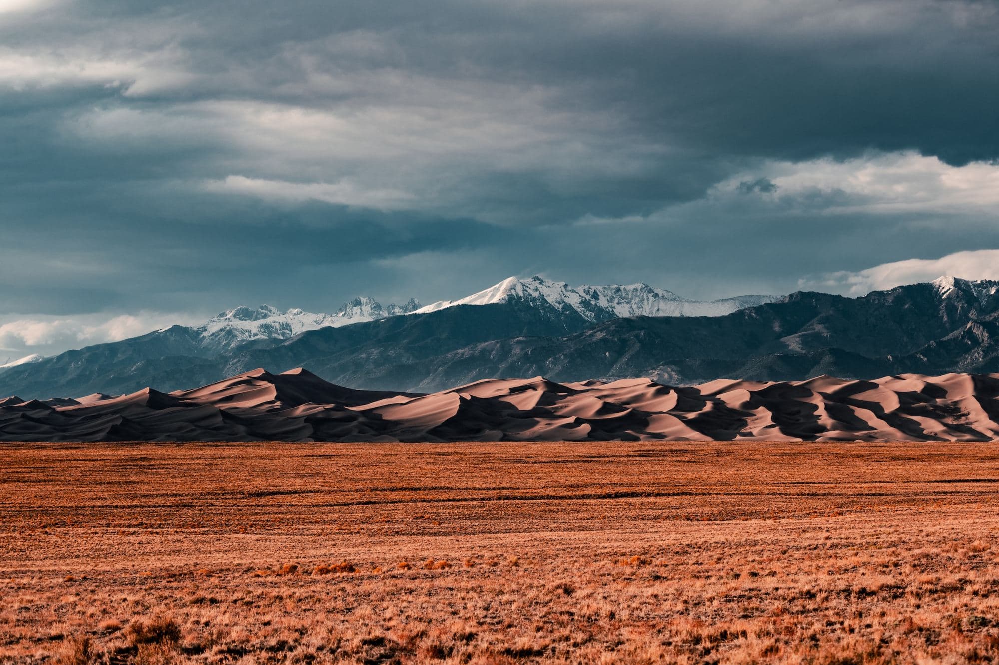 The Great Sand Dunes of Colorado from across a dim-lit field with snow-capped mountains in the far distance and heavy cloud cover (photo by Colin Lloyd)