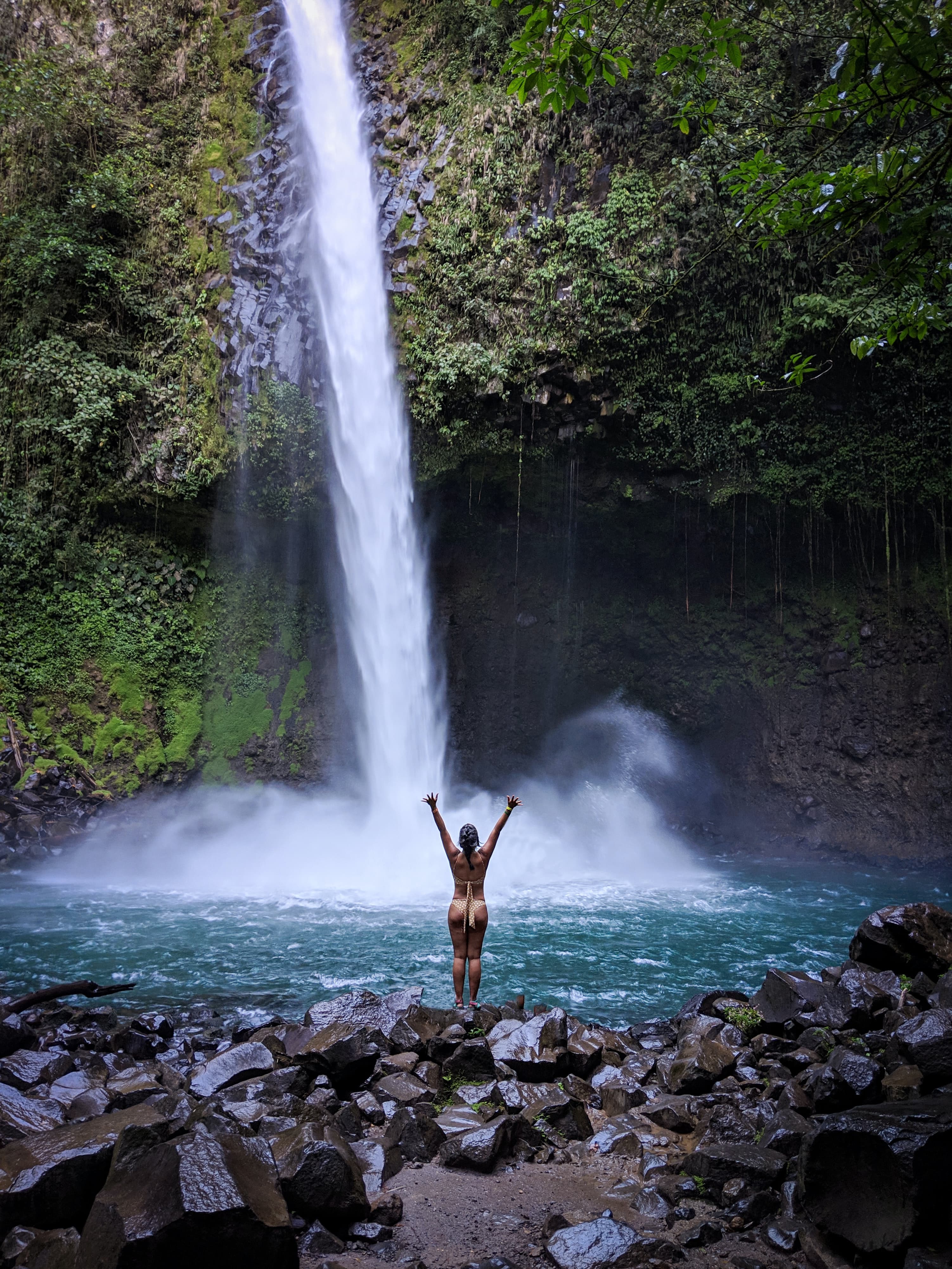 girl standing in front of waterfall surrounded by green trees