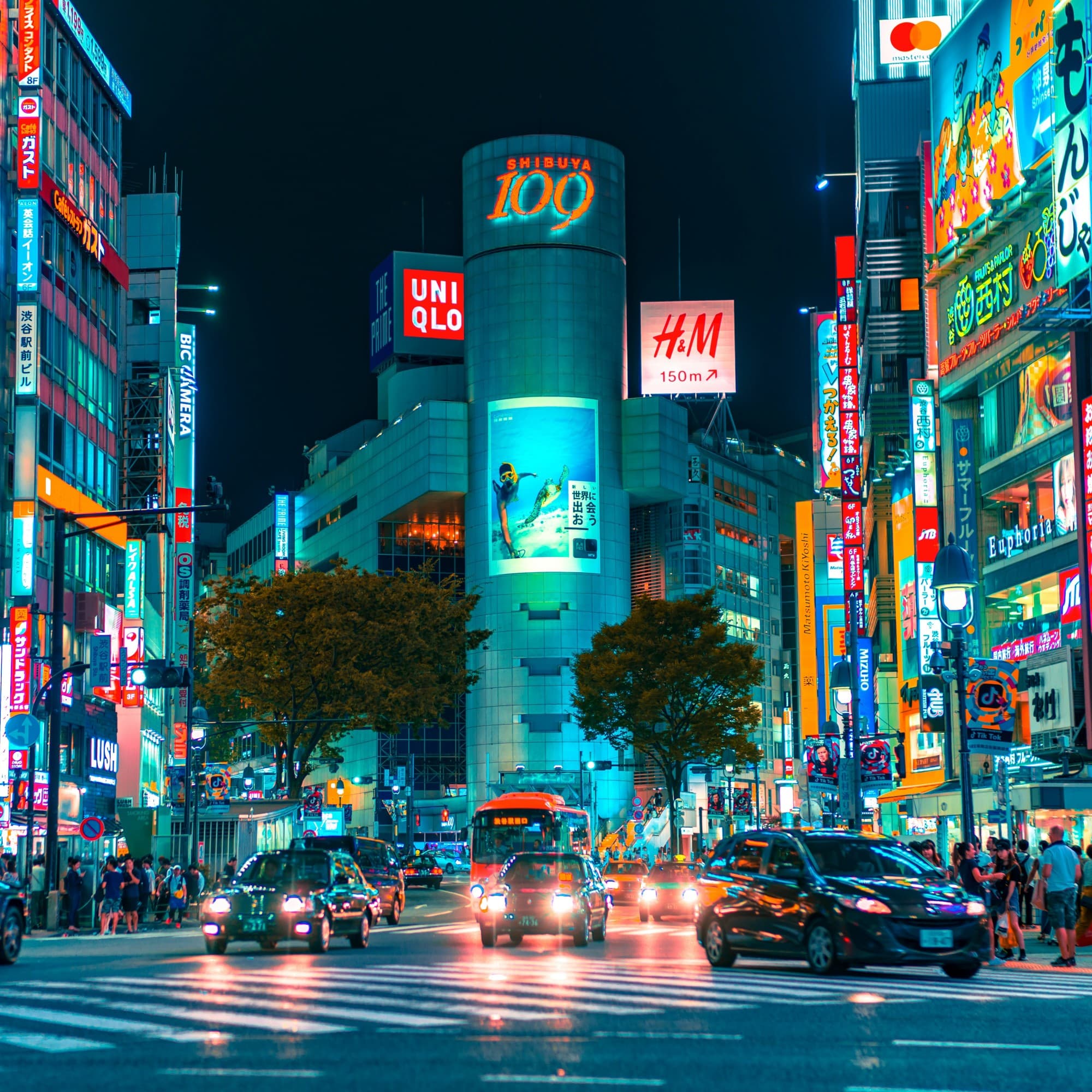 A view of city buildings lit up at night in Japan surrounded by cars and tourists.