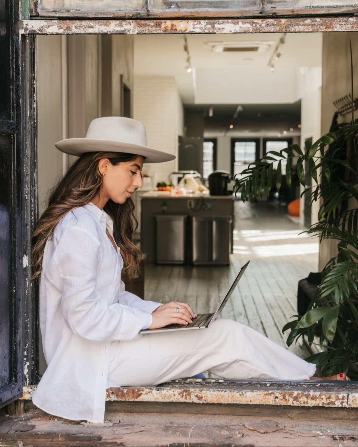 a woman in a brimmed hat sits on a windowsill and works on her laptop