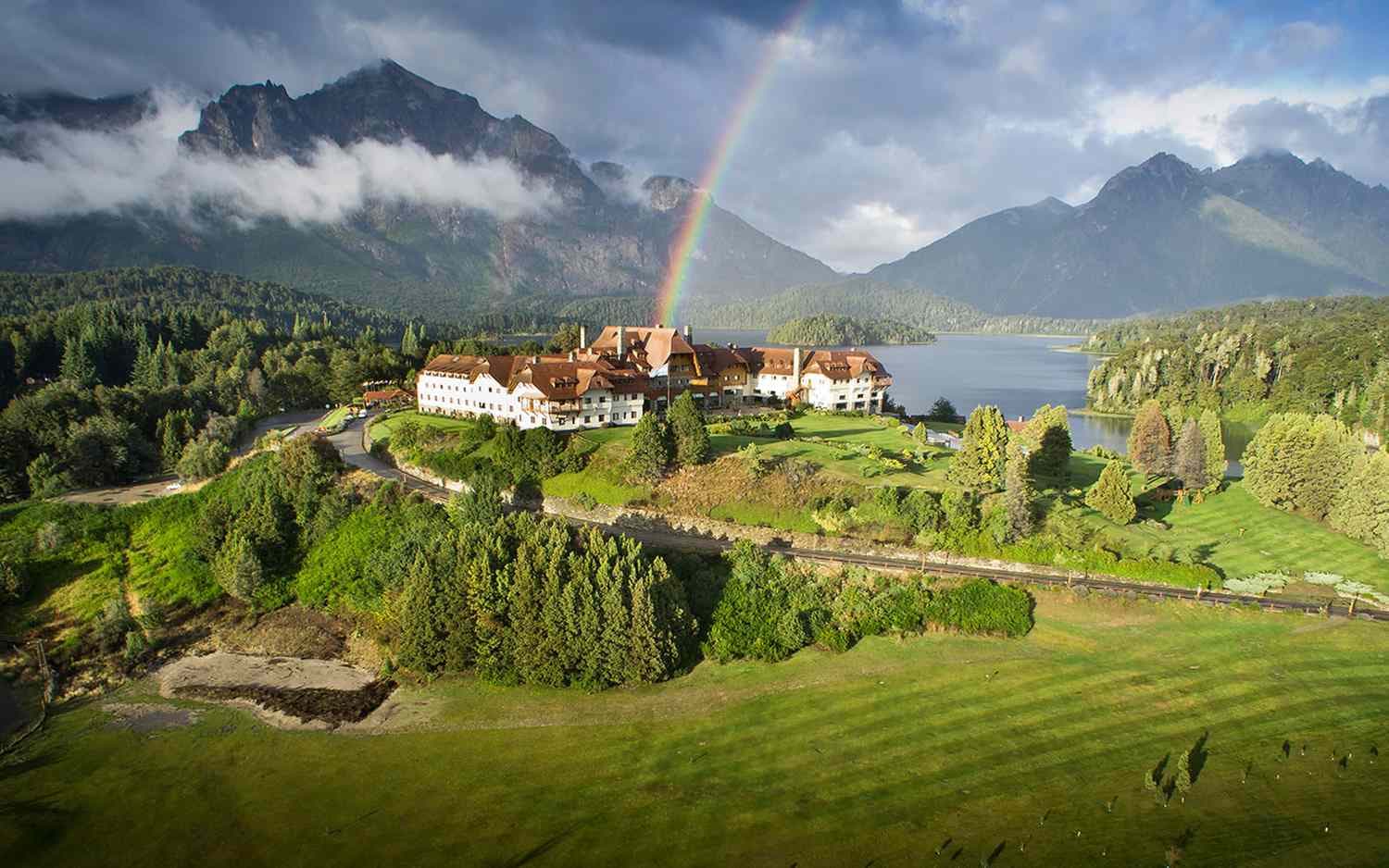 Beautiful scenery with a large estate on a hill by a lake and mountains with a rainbow overhead.