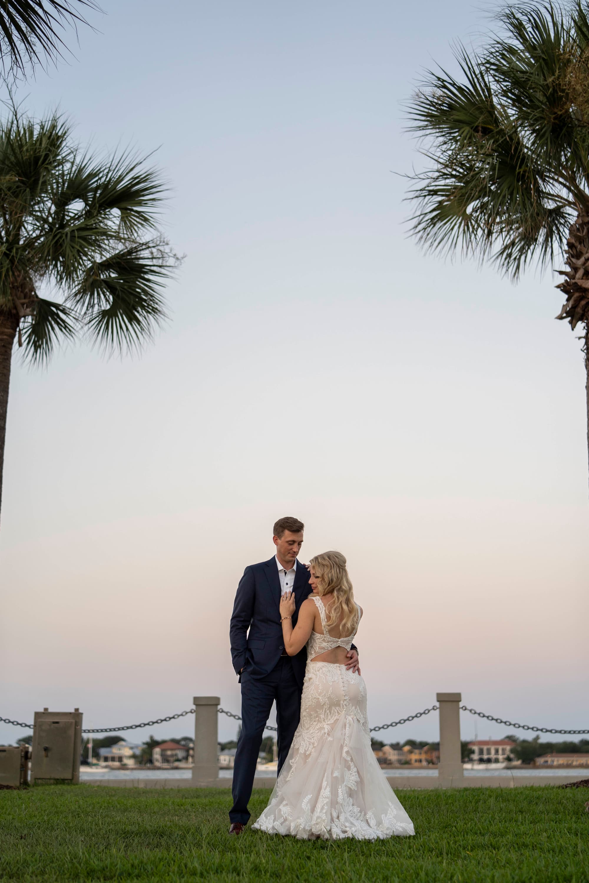 Two individuals in formal attire stand facing each other between palm trees at twilight.