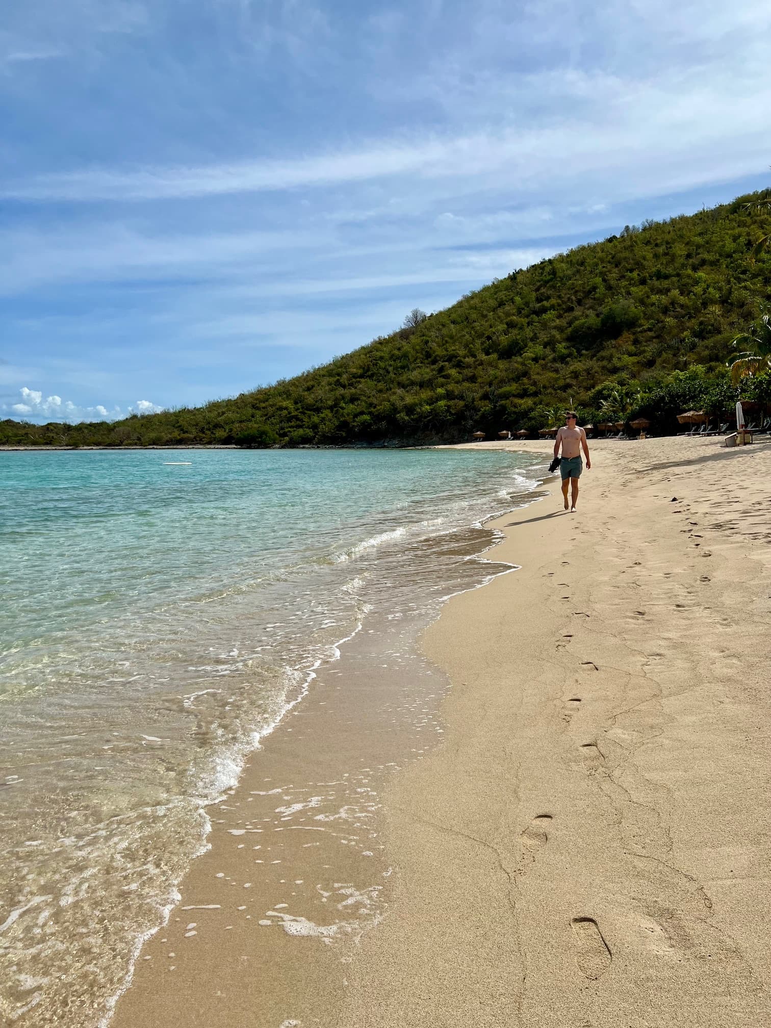 A man walking down a beach with crystal clear water and a green mountain in the background.